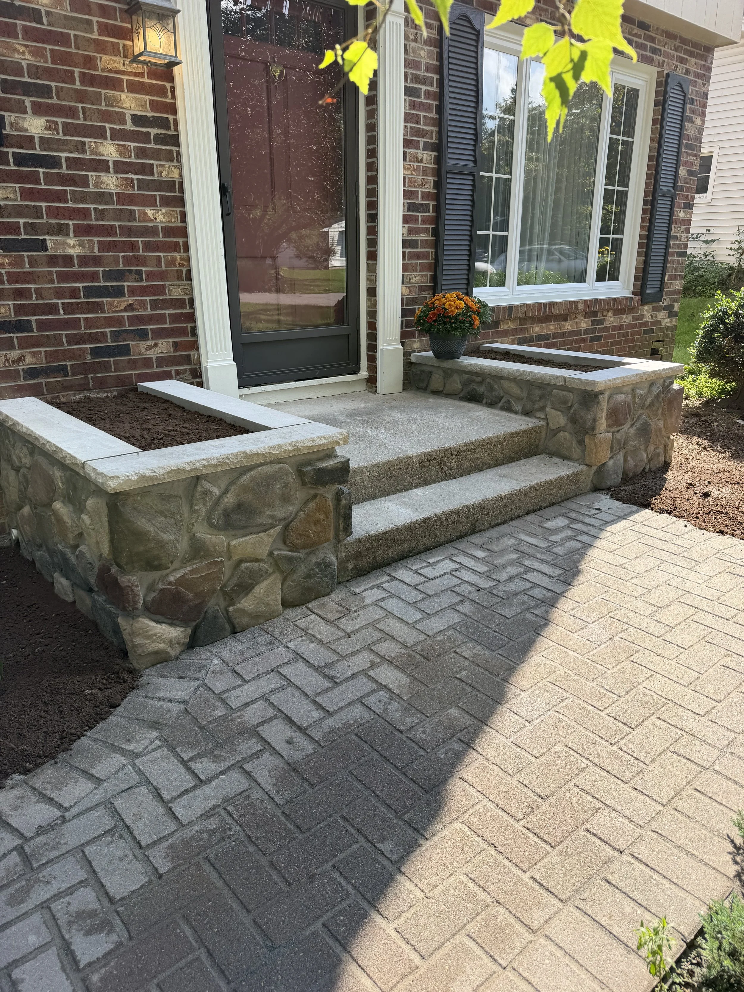 Front porch with brick house, white framed window with black shutters, black door with screen, outdoor light, flower pot with orange chrysanthemums, stone border, and a brick and concrete walkway.