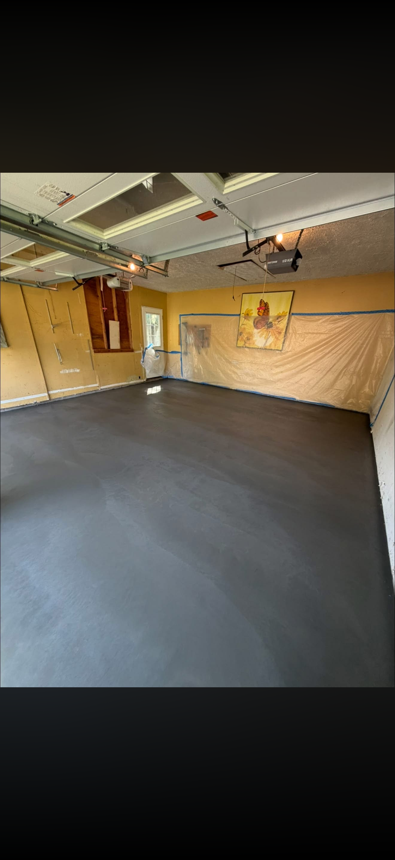 A garage with a newly poured concrete floor, yellow walls, a window, and a partially finished ceiling. The garage door is open, revealing the ceiling mechanics, and a colorful painting hangs on the wall, covered partially with protective plastic.