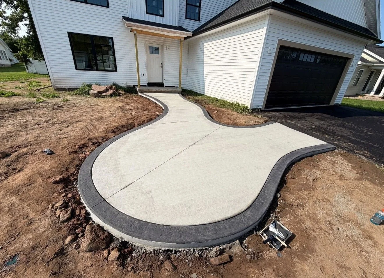 New concrete sidewalk with curved design leading to front door of a white house, adjacent to a black garage door, with the yard still under construction and no grass yet.