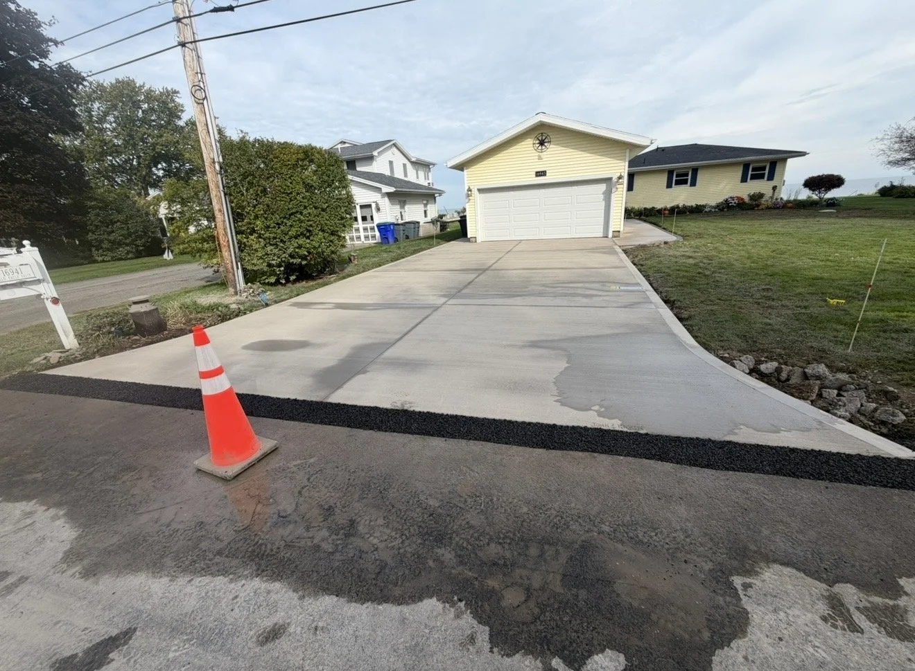 Newly paved concrete driveway in front of a yellow garage with a decorative compass rose above the garage door, leading to a house and garden, with traffic cone and construction markings nearby.