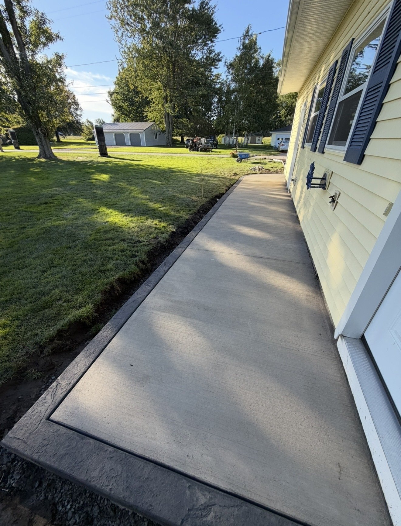Newly installed concrete sidewalk in front of a yellow house with blue shutters, with grass and trees in the background under a clear sky.