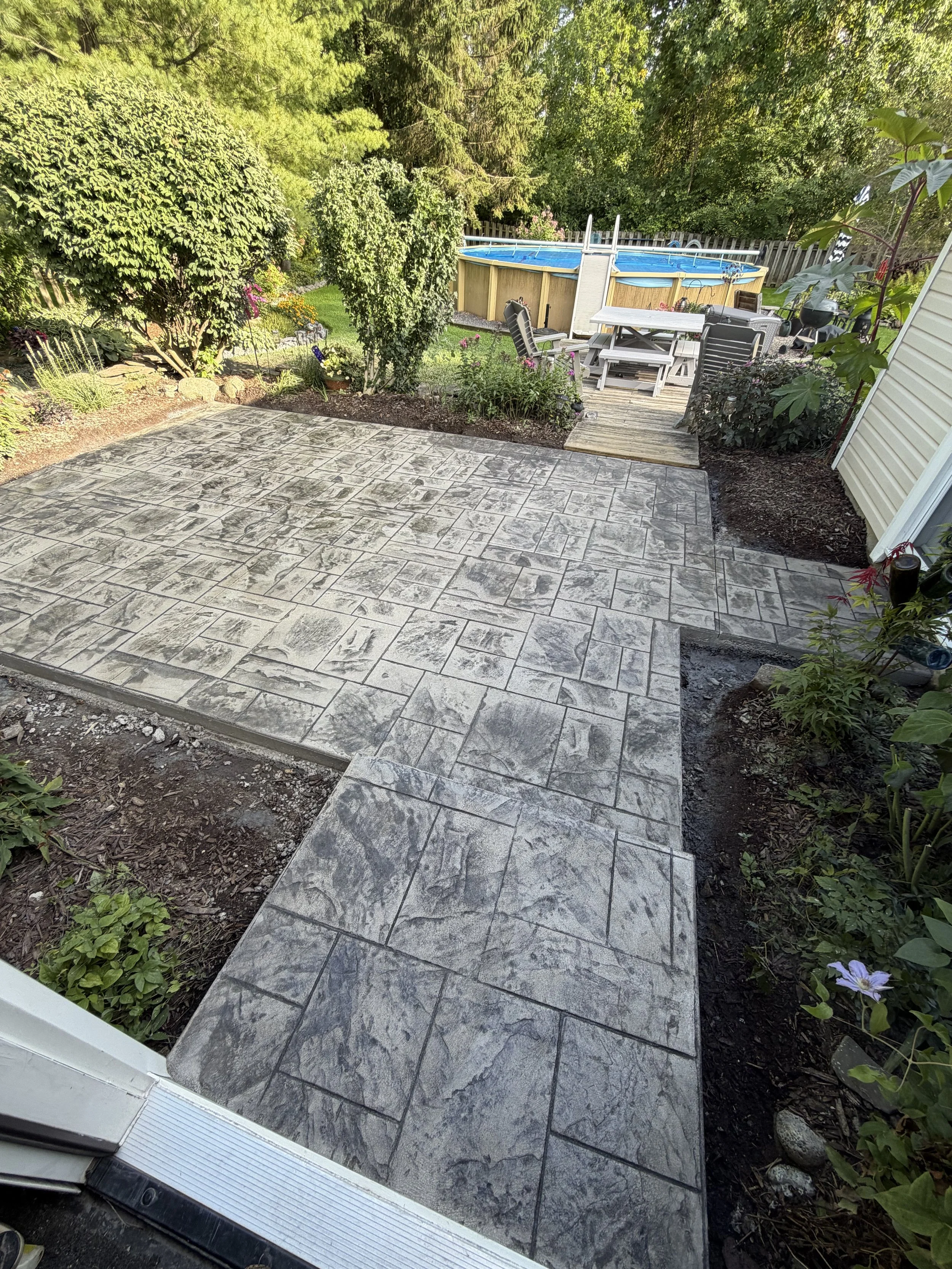 Backyard patio with stamped concrete, garden beds, trees, a deck with outdoor furniture, a above-ground swimming pool, and fencing.