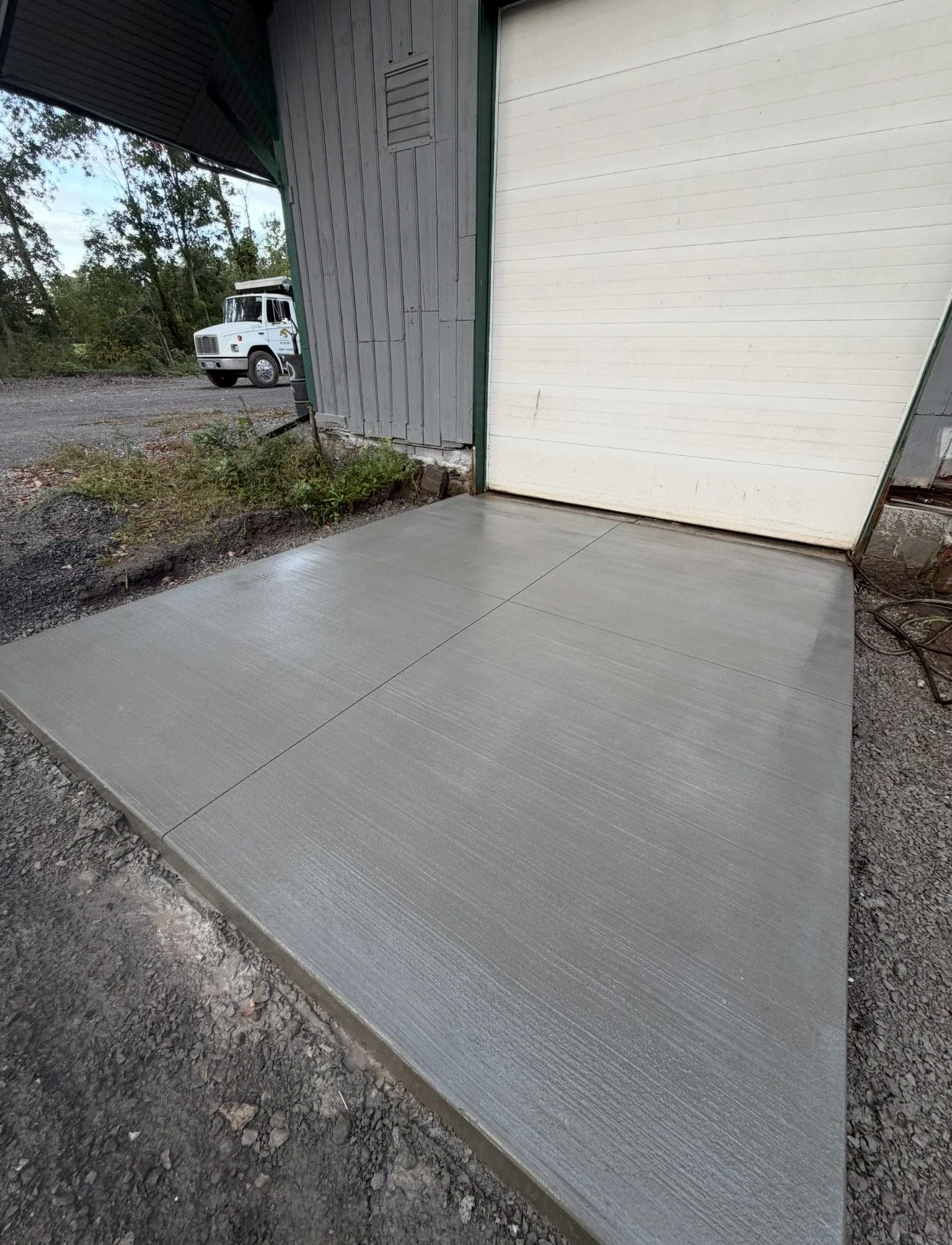 Newly poured concrete driveway in front of a garage, with a closed white garage door, in a rural area. A white delivery truck is visible in the background on a gravel area, with trees and blue sky behind it.