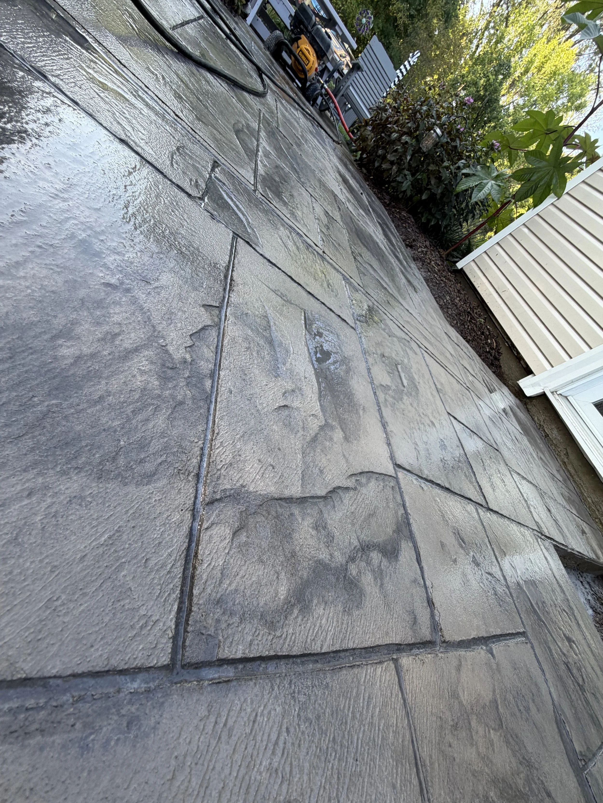 A close-up view of a newly cleaned gray stone patio with visible water sheen, surrounding a house with siding, plants, and outdoor tools in the background.