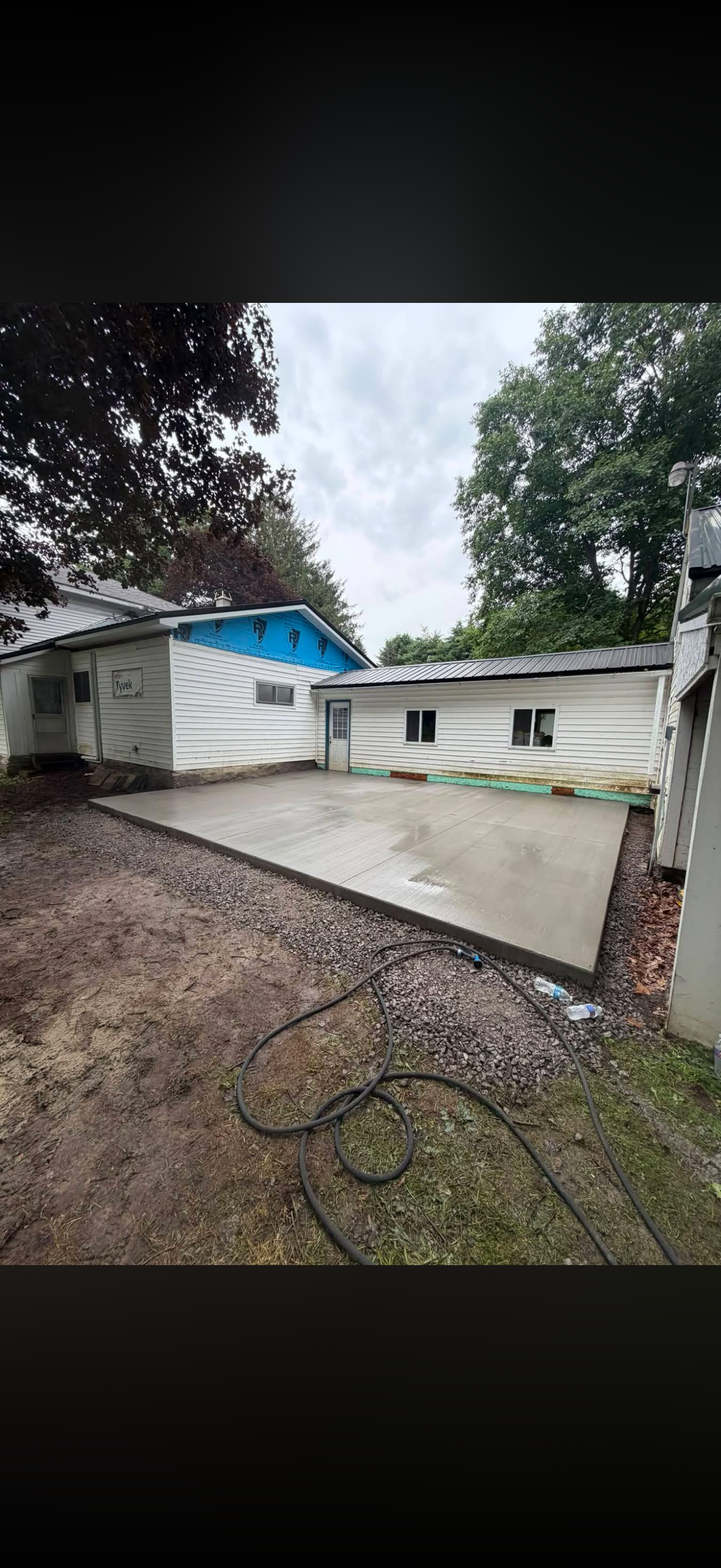 Newly poured concrete patio in backyard with water on surface, adjacent to a white house with some construction work happening, overcast sky, trees in background.