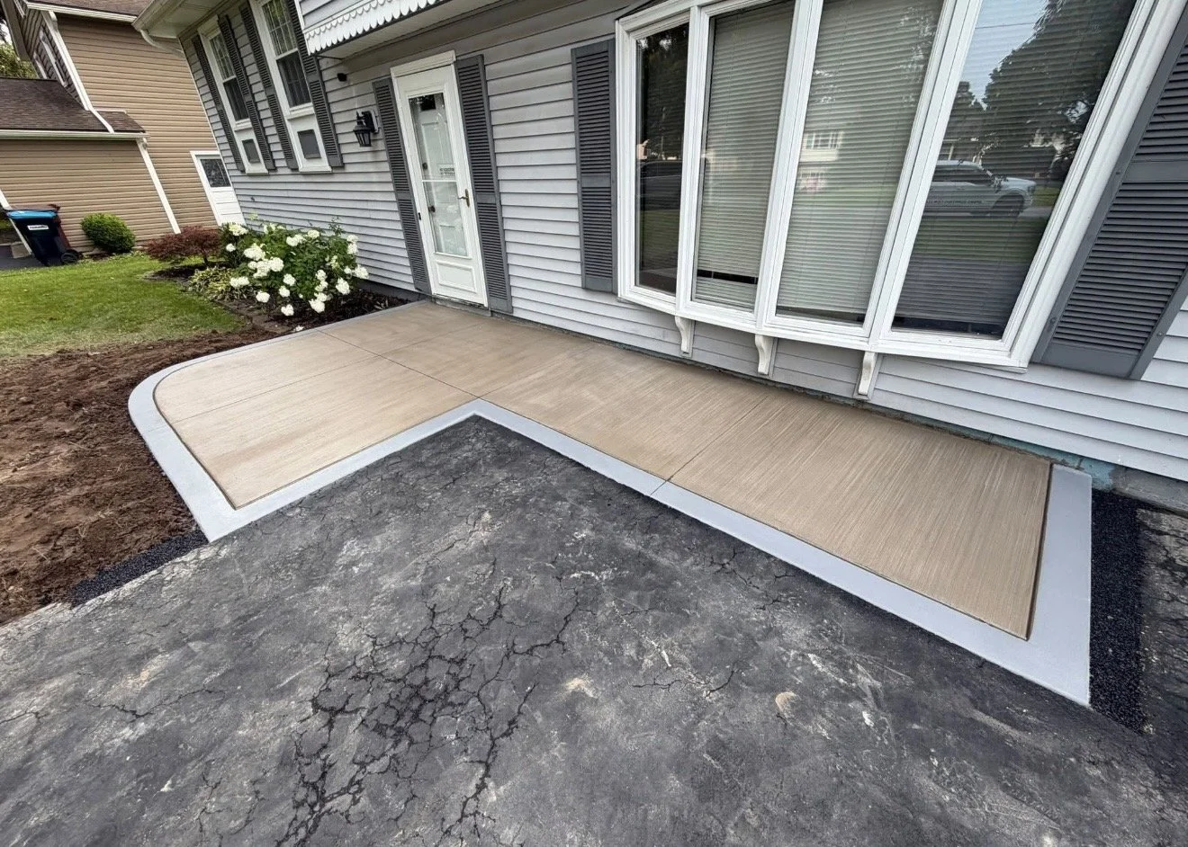Newly installed concrete steps with a textured beige surface lead to the house door and window, with a flower bed of white hydrangeas nearby and a cracked asphalt driveway in the foreground.