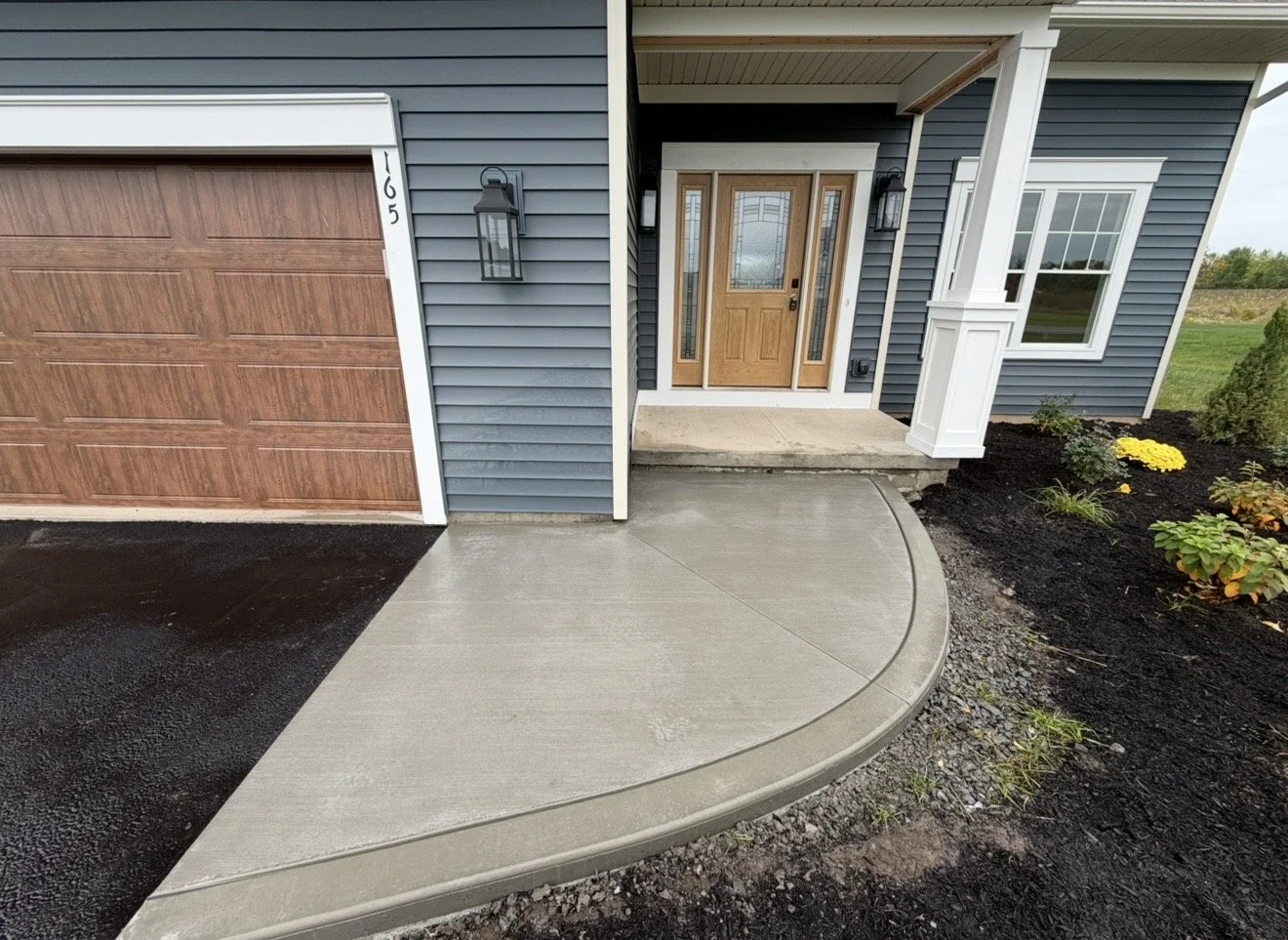 Front entrance of a house with a wooden door, a small porch, a concrete driveway, a garden bed with yellow flowers, and attached garage with brown door.