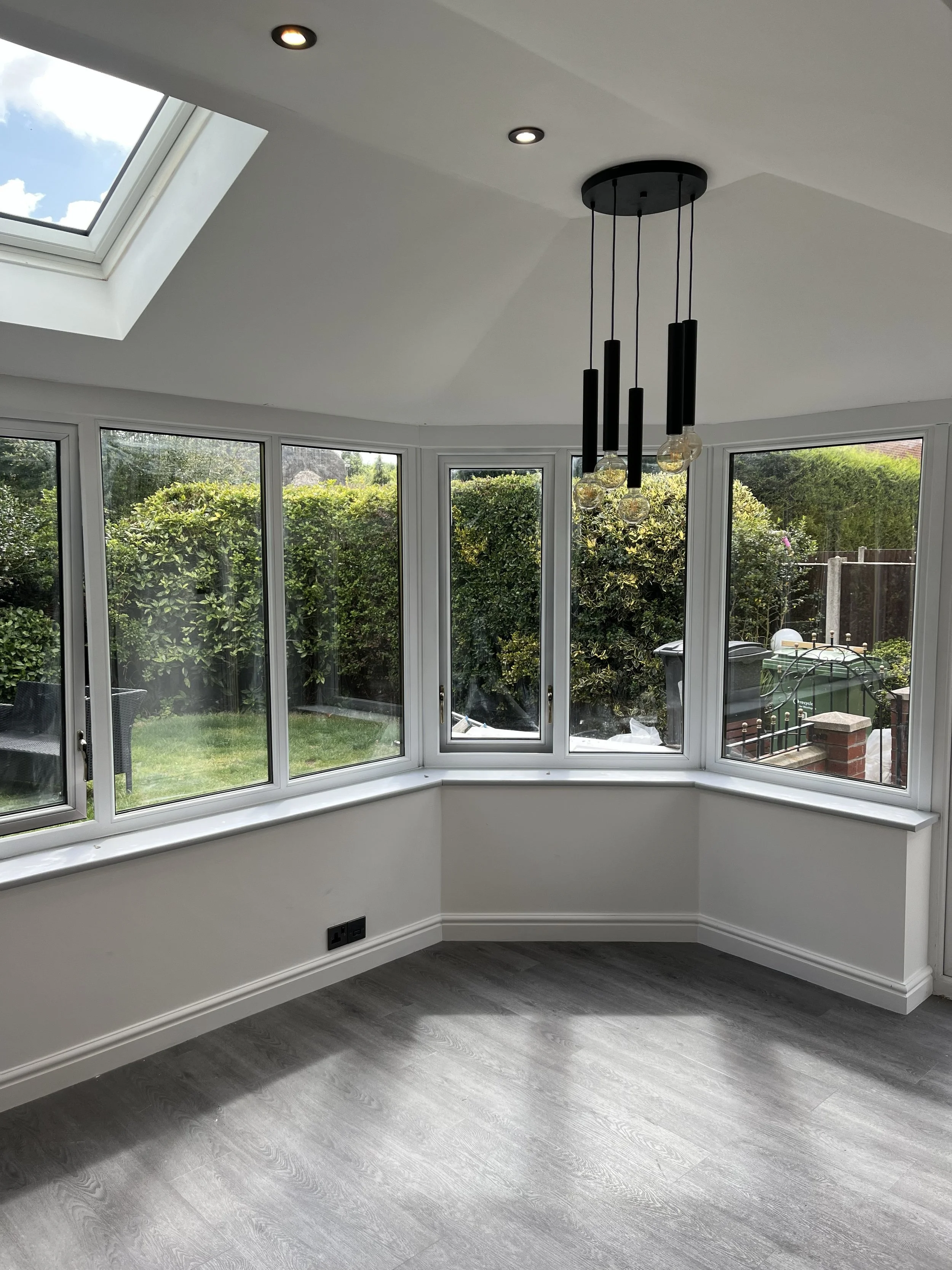 A bright sunroom with large windows and a skylight, featuring a modern black chandelier with multiple hanging bulbs, white walls, and gray wood flooring, overlooking a green backyard with outdoor furniture.