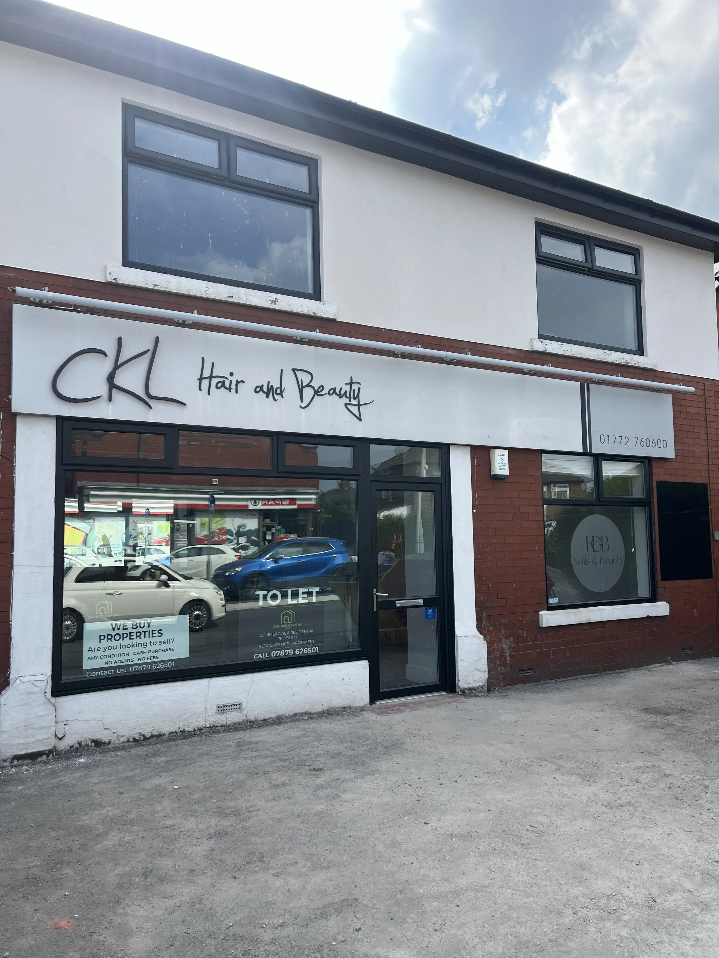 A two-story building with a hair and beauty salon on the ground floor. The sign reads 'CKL Hair and Beauty.' The windows are dark and reflective, with a large window in the front and smaller ones on the second floor. There is a 'TO LET' sign on the glass door and a parking lot with some cars in front. The sky is partly cloudy.