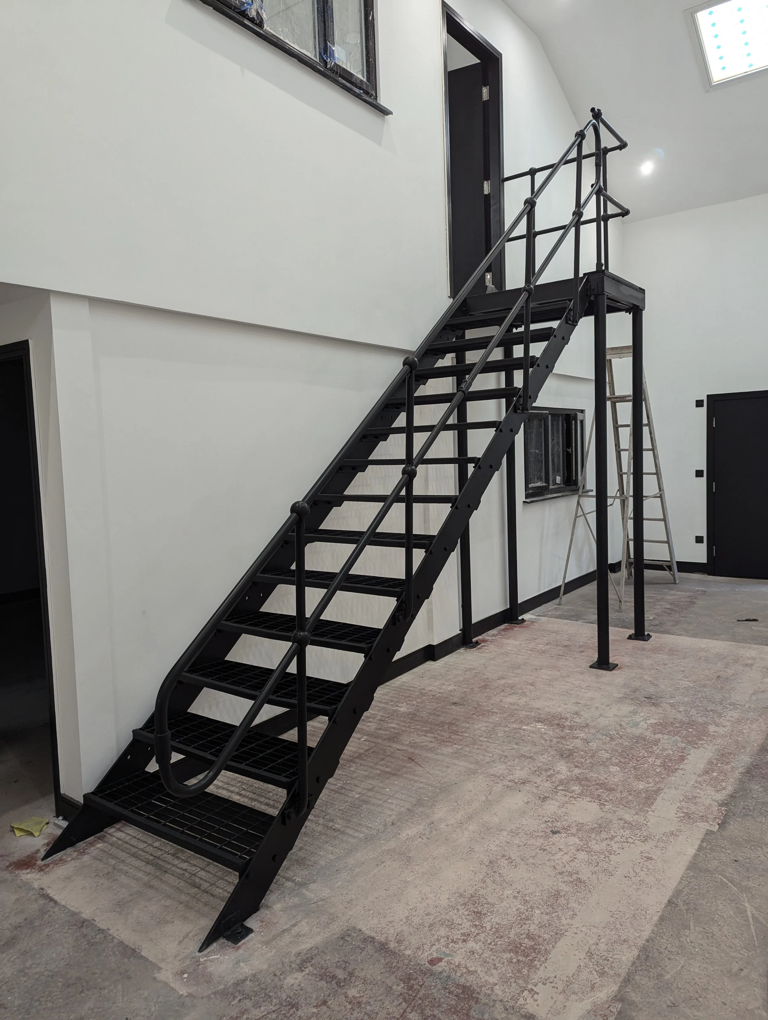 Interior of a building under construction or renovation, with black metal staircase and a ladder next to it. The walls are painted white, and there is a window with blue protective film on the upper part of the wall. The floor appears to be unfinished or partially covered.