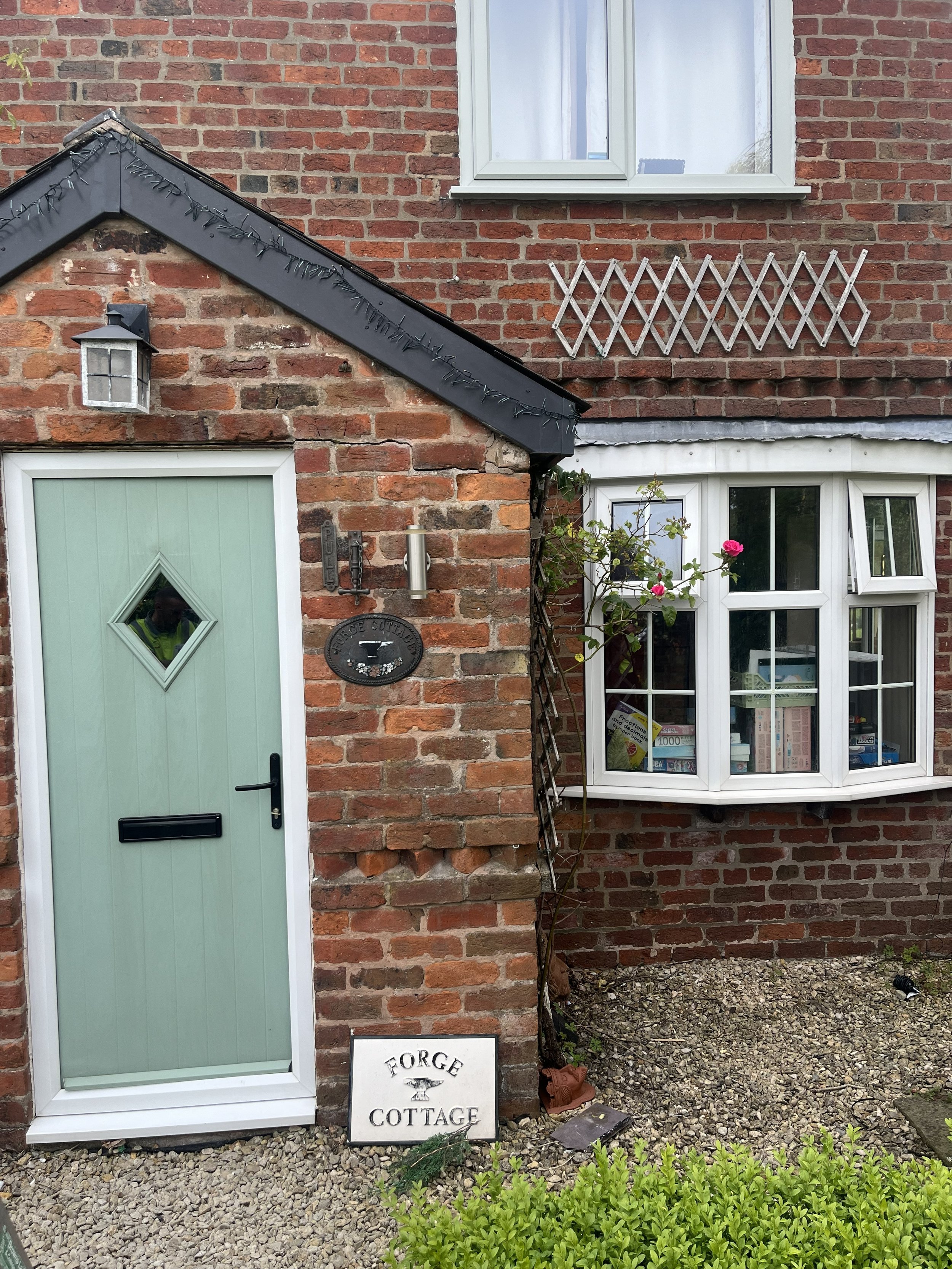 The image shows the front of a brick house with a green front door with diamond-shaped window panes, a hanging lantern light, and a rectangular sign reading 'Forge Cottage' near the ground. There is a bay window with white-framed panes and several books and decorations inside. An open window with a white frame is above the bay window. A small plant with pink flowers climbs next to the window. The ground is covered with gravel, and there are bushes in the foreground.