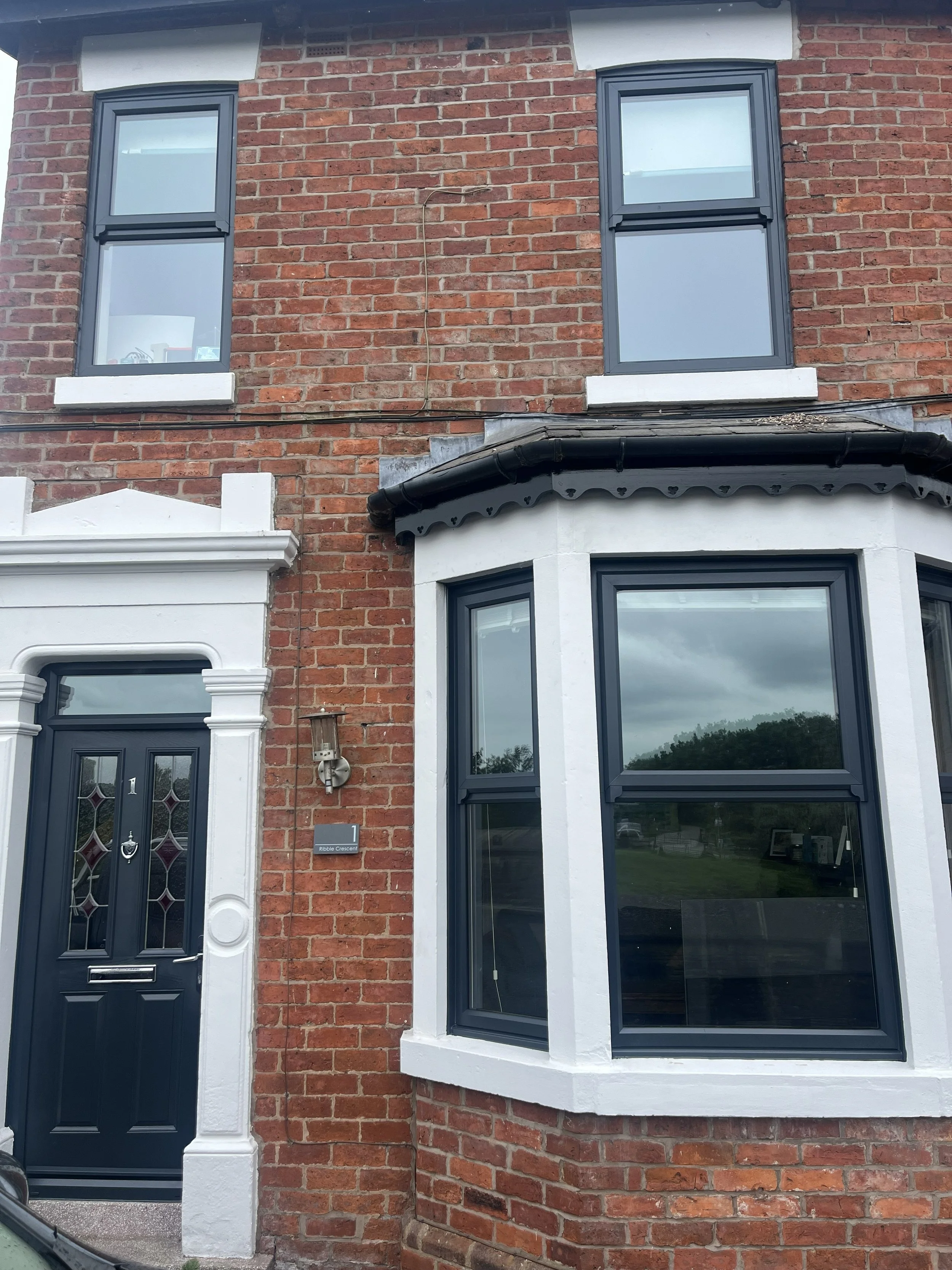 Front of a brick house with a black door featuring decorative glass, a white door frame, and a white column on the left, with two large windows with black frames, one on the upper level and one below. The house has a black gutter and a small area of grass reflected in the window.