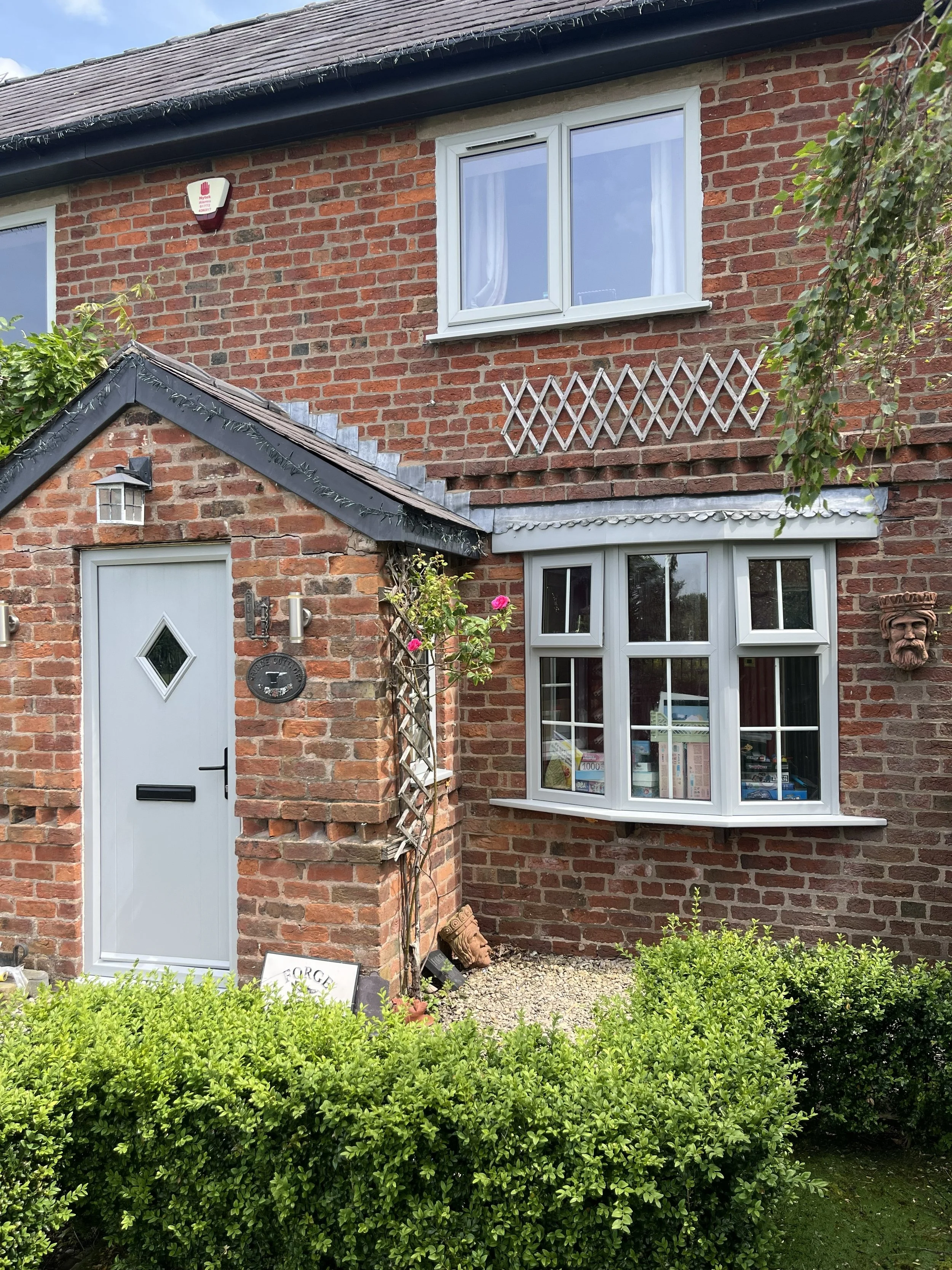 Brick house with a white front door, a large window with white trim, and a small window above a garden with green bushes. Decorative wooden lattice and a small sculpture or face on the wall are visible.