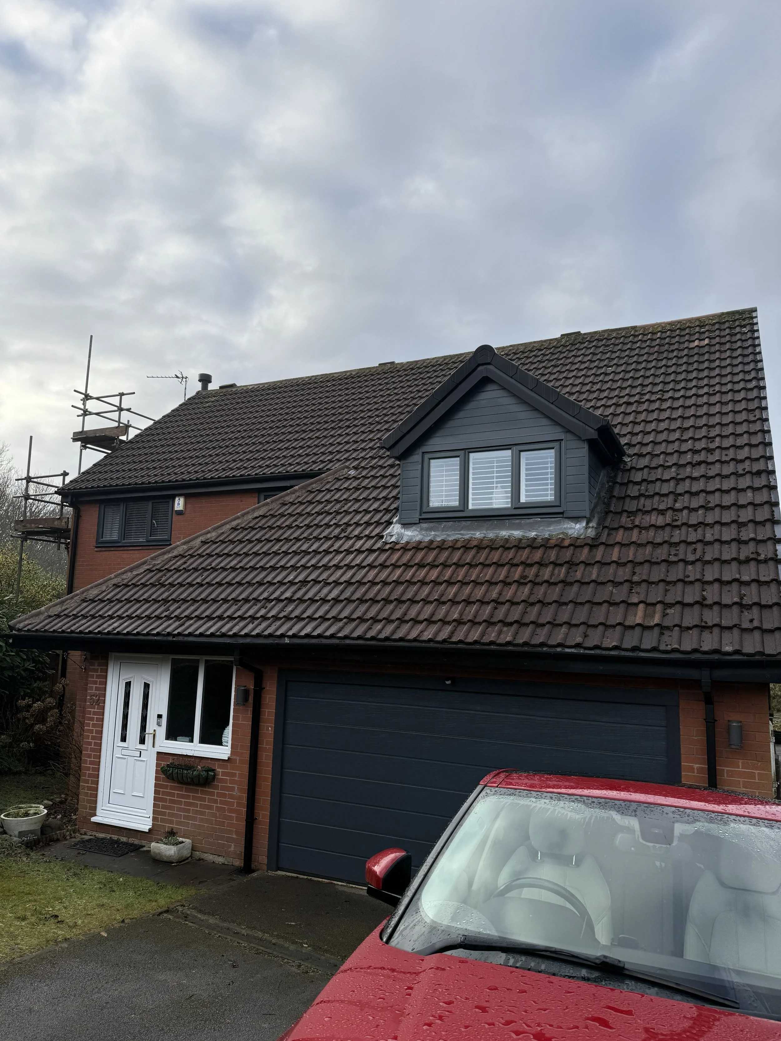 Front of a brick house with a double garage, white front door, and an attic window. A red car is parked in front, and the house has a tiled roof with a dormer window. The sky is cloudy.