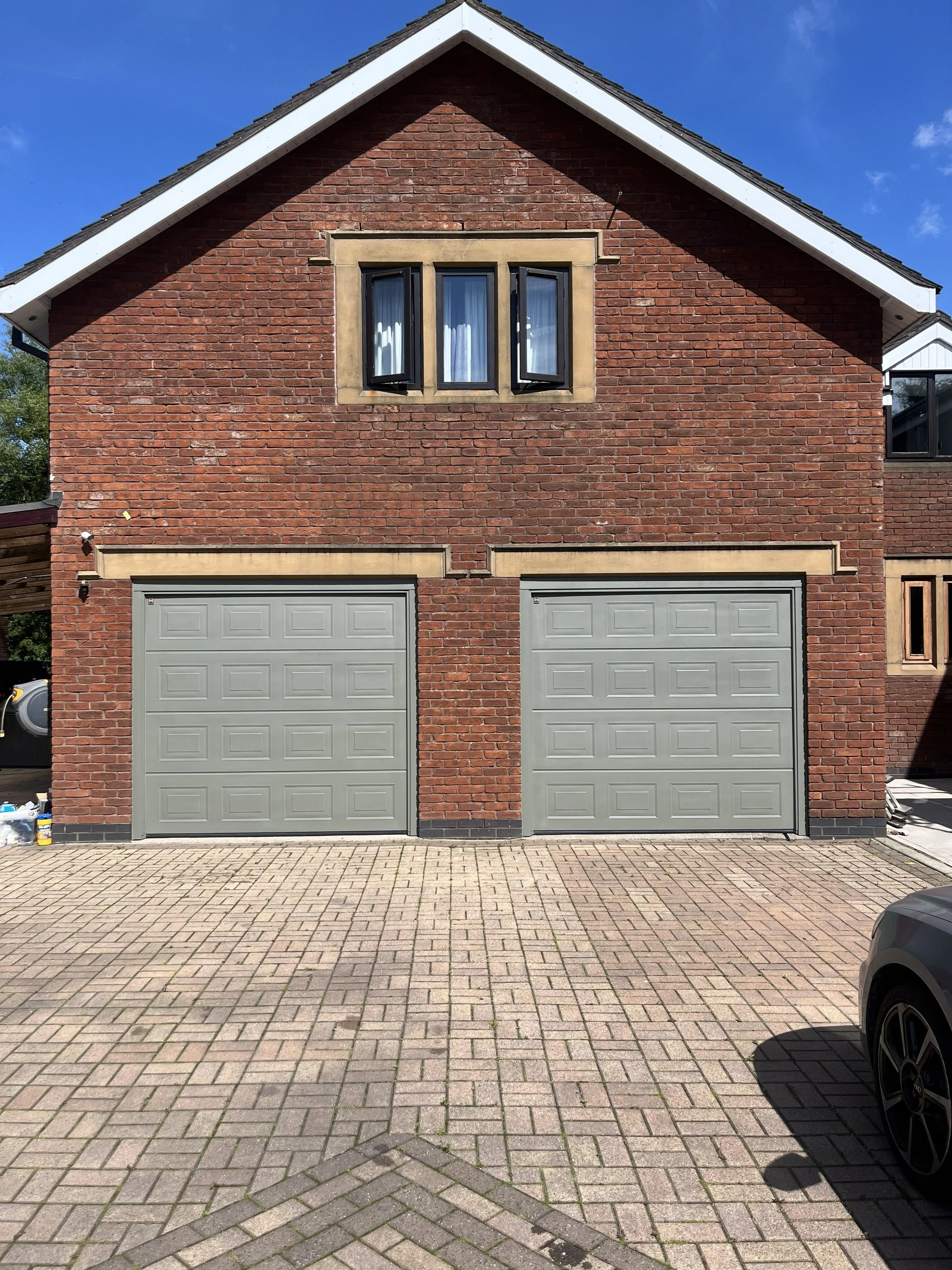 Red brick house with two gray garage doors and a window above them. The house has a gable roof with dark shingles and white trim, and is situated in a paved driveway with interlocking bricks.