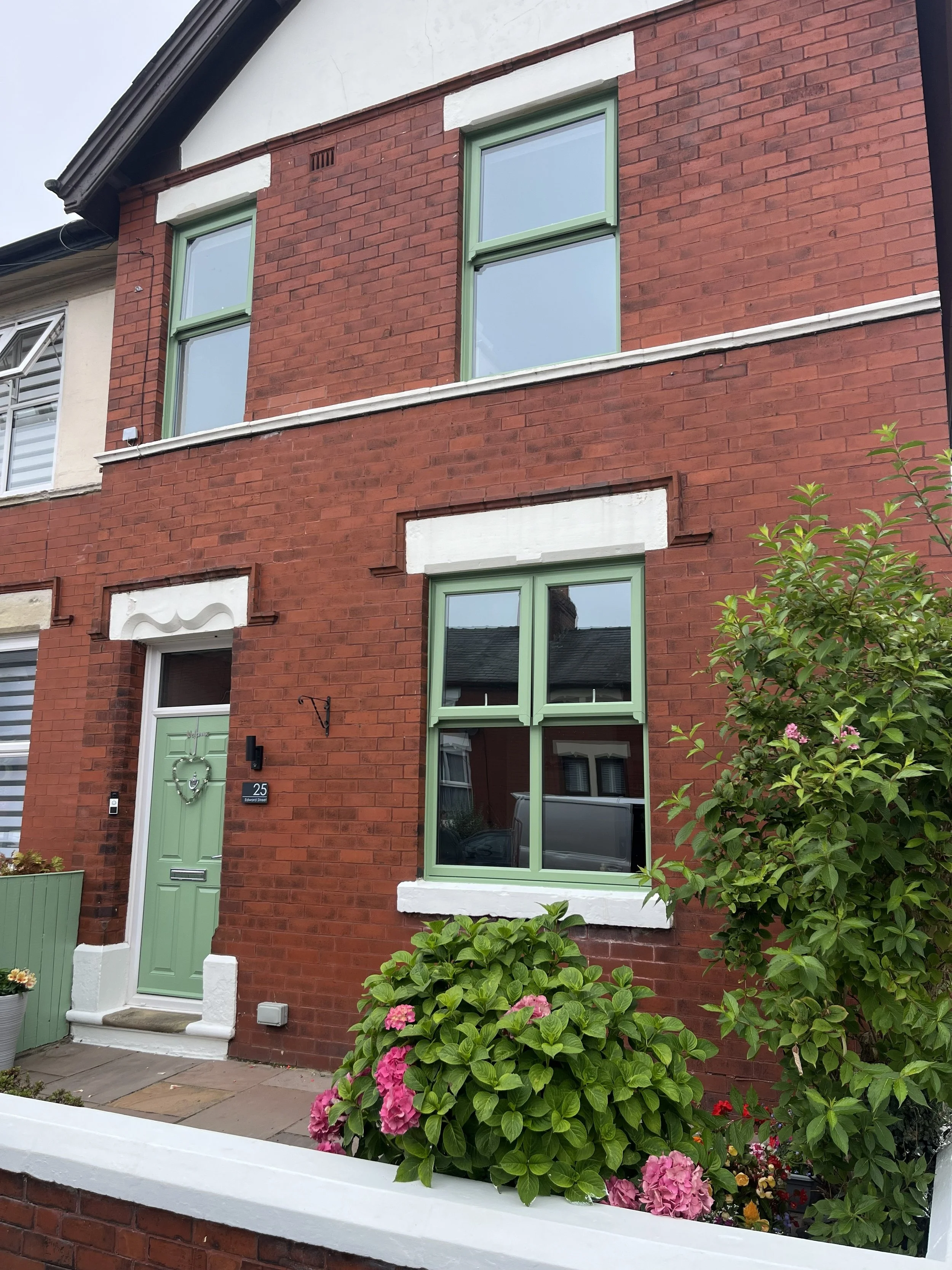 A red brick house with painted green window frames and a matching front door. There are pink hydrangea bushes in the garden in front of the house.