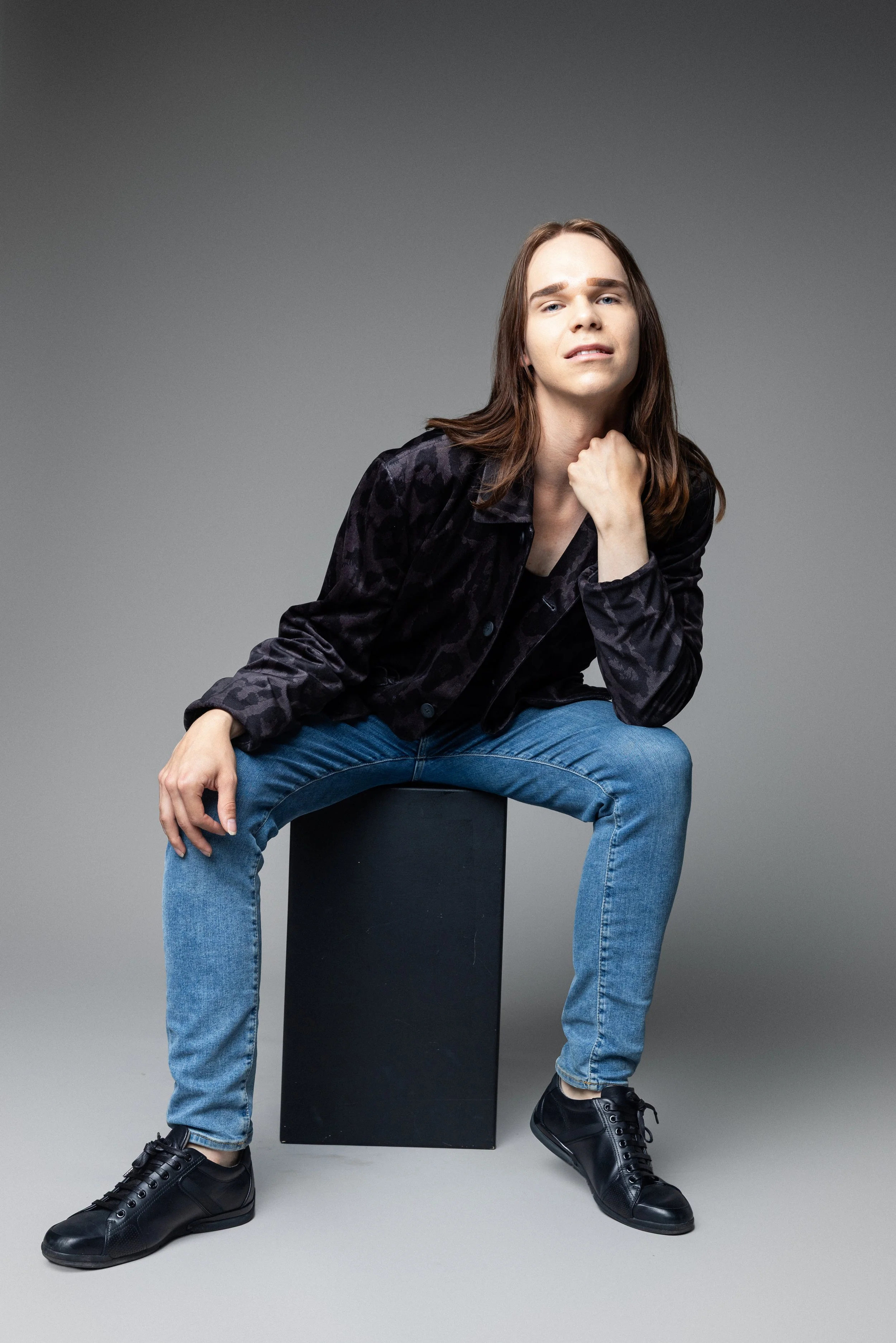 A woman with long brown hair, wearing a dark patterned jacket, blue jeans, and black sneakers, sitting on a black block against a gray background.