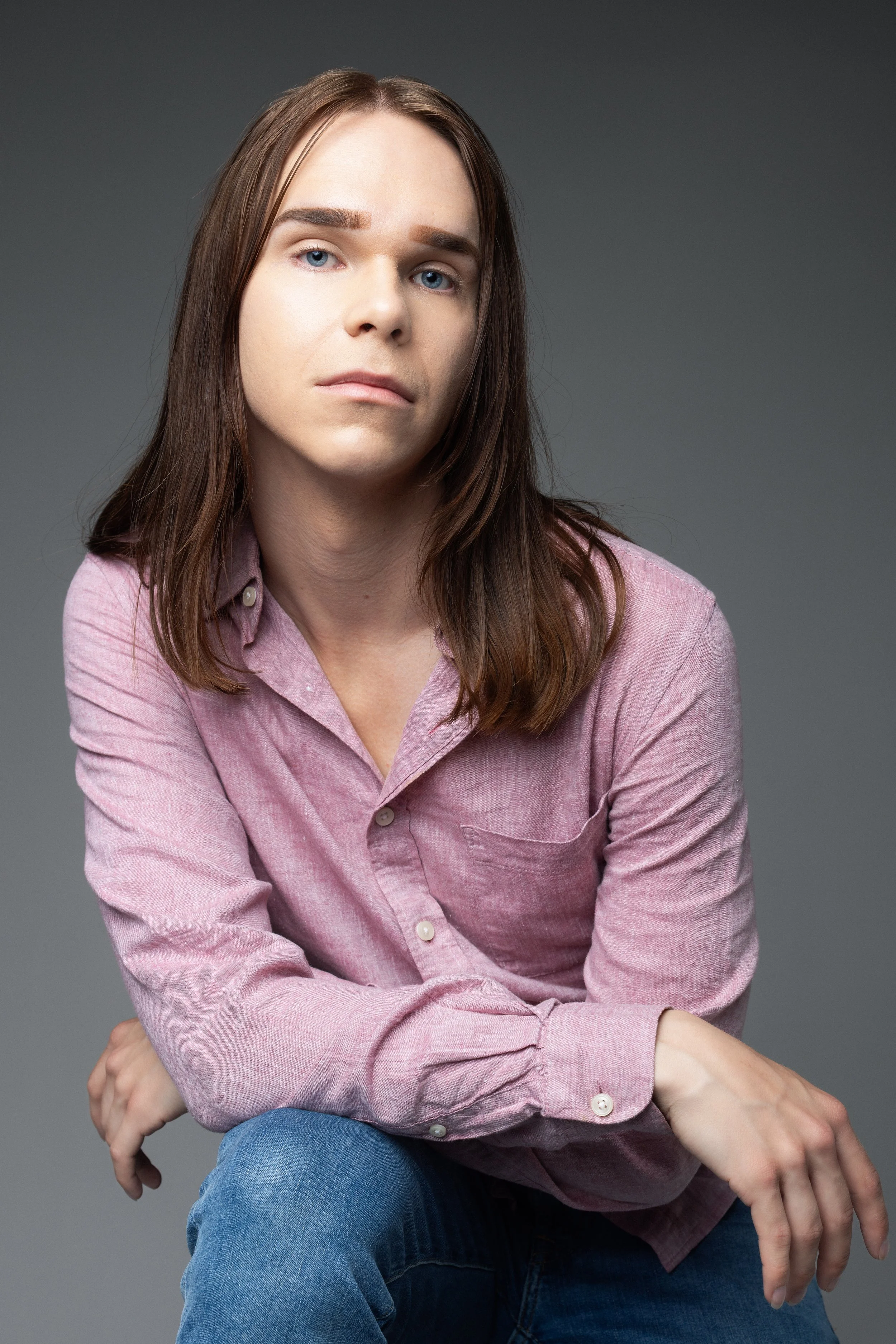 Portrait of a person with long brown hair, wearing a pink button-up shirt and blue jeans, sitting against a plain gray background.
