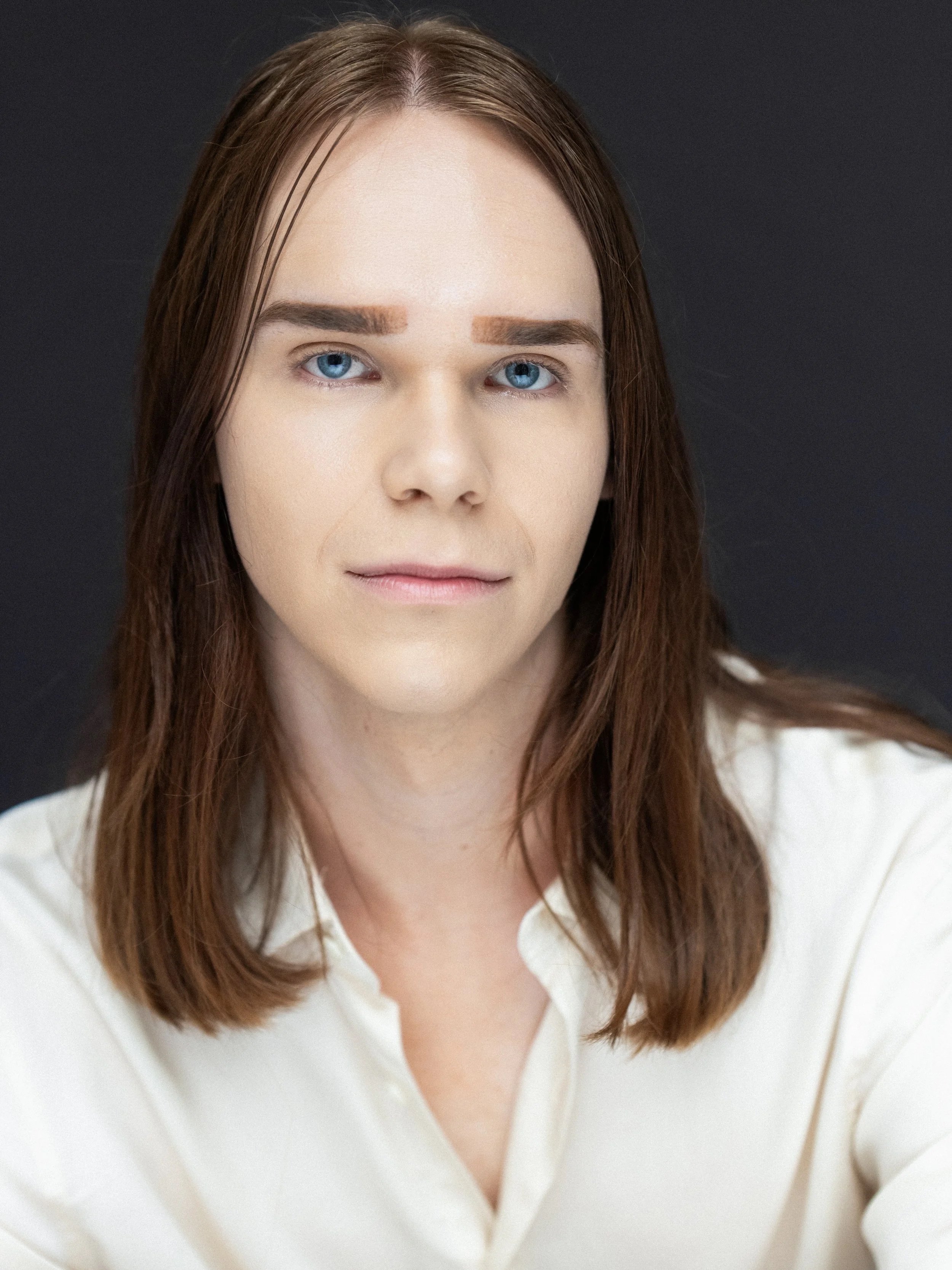 A woman with long brown hair and blue eyes wearing a white shirt against a dark background.