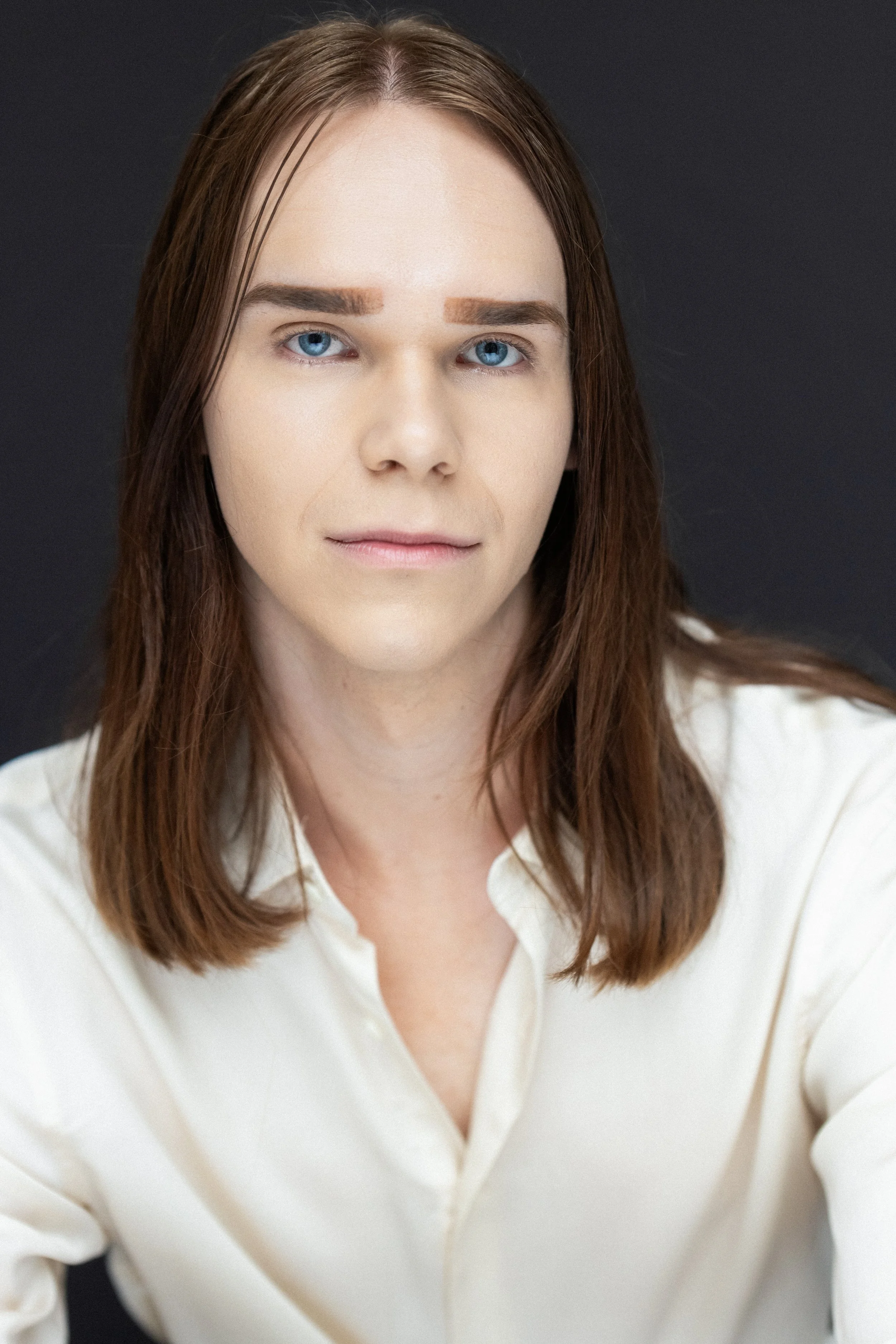 A woman with brown hair, light skin, and blue eyes wearing a white shirt and looking at the camera, set against a dark background.
