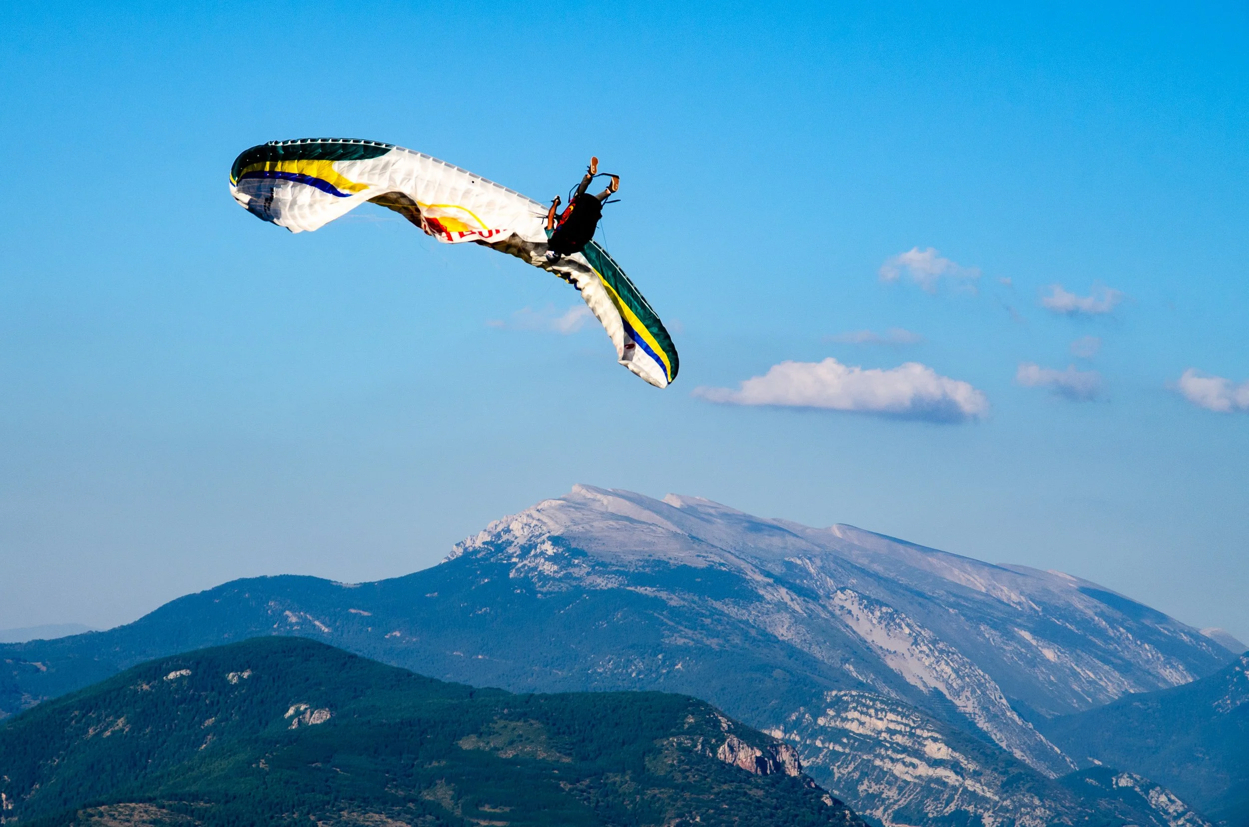 A person paragliding over mountains with snow-capped peaks and green forests, under a clear blue sky.
