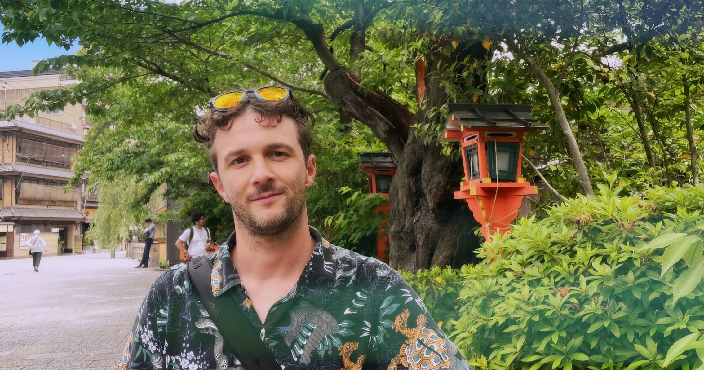 A young man with brown curly hair and sunglasses on his head stands in front of a lush green tree with orange and black traditional Japanese lanterns, in an outdoor urban setting with other people walking by.