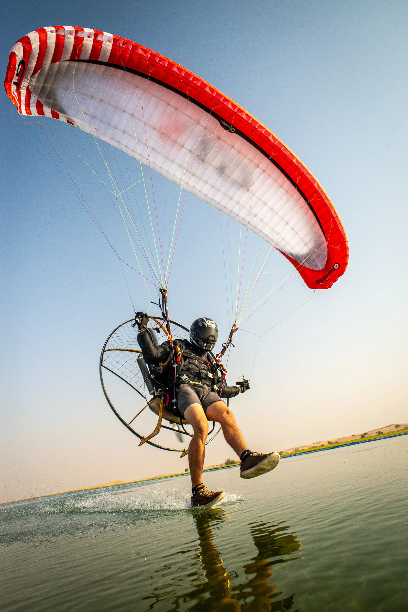 A person in a powered paraglider flying low over a body of water with a clear sky in the background.