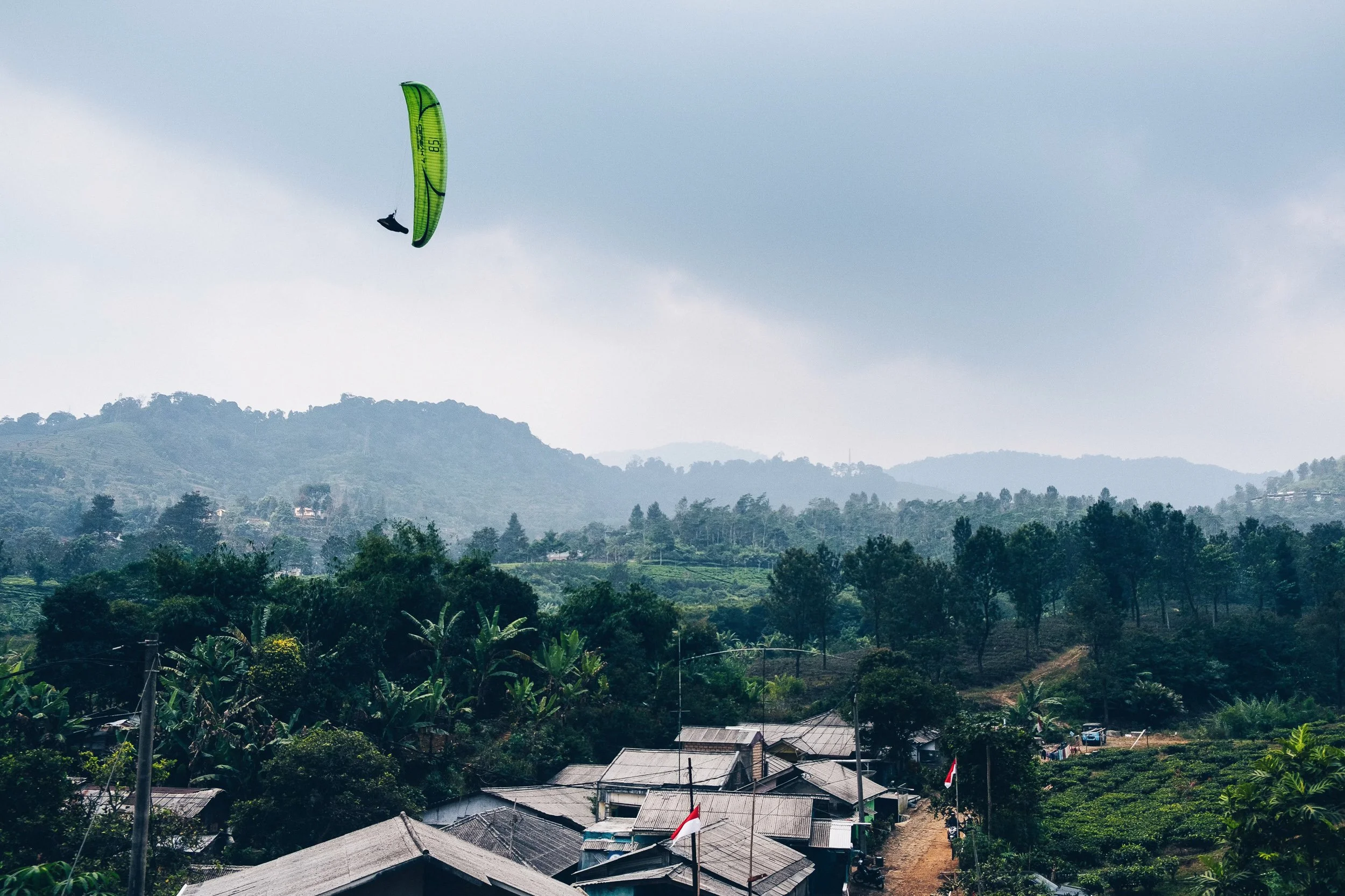 Scenic view of a rural landscape with small houses, trees, and greenery in the foreground, rolling hills, and mountains in the background under a cloudy sky. A person is flying a green kite high in the sky.