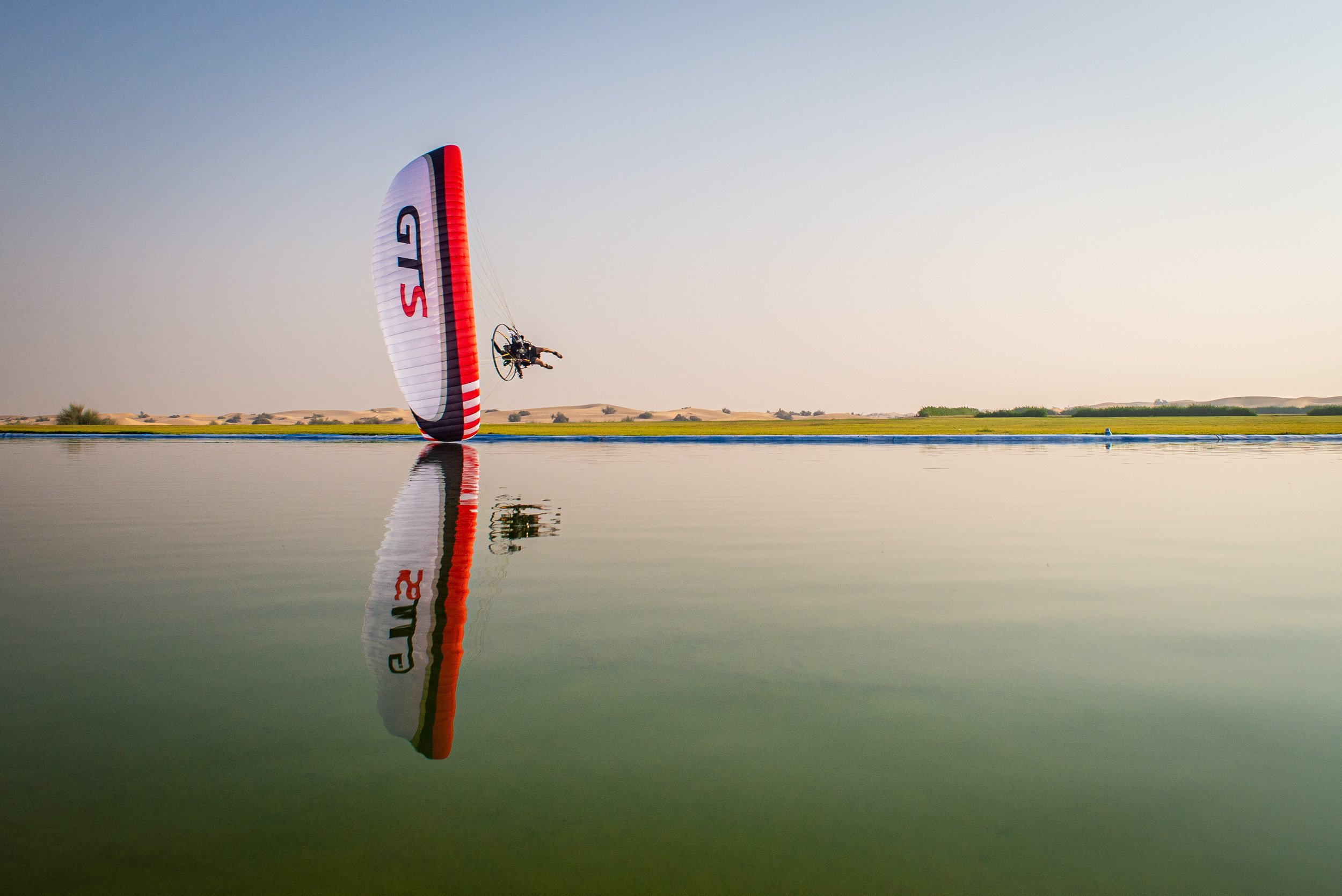 A powered paraglider flying over a body of water with a grassy landscape and sand dunes in the background.