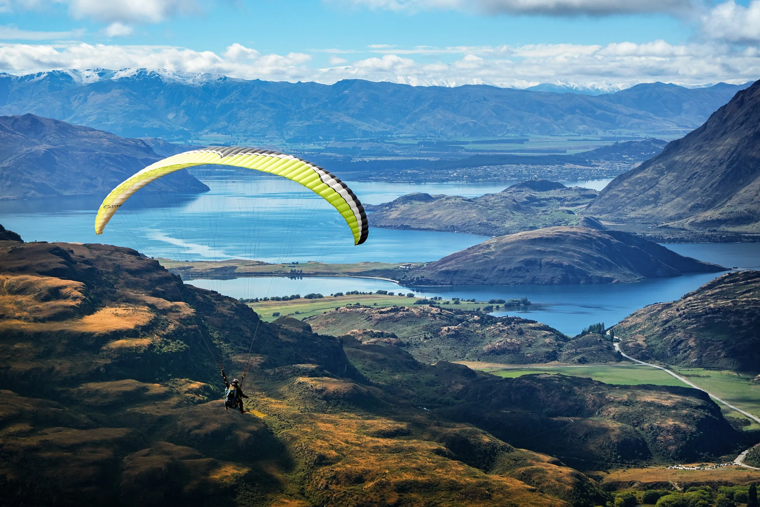 Paraglider flying over a mountainous landscape with lakes, valleys, and snow-capped peaks in the distance.