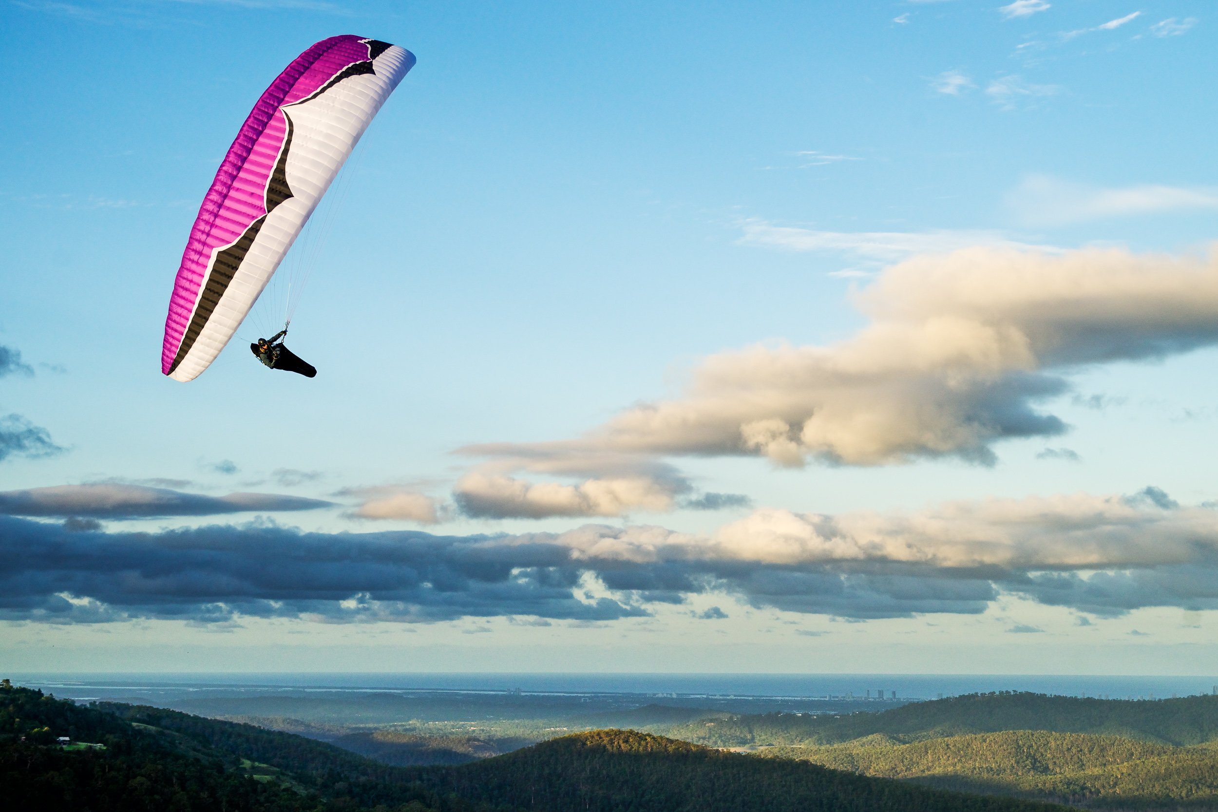 A person paragliding with a pink, white, and black parachute over a scenic landscape of rolling hills, water, and distant city skyline under a partly cloudy sky.