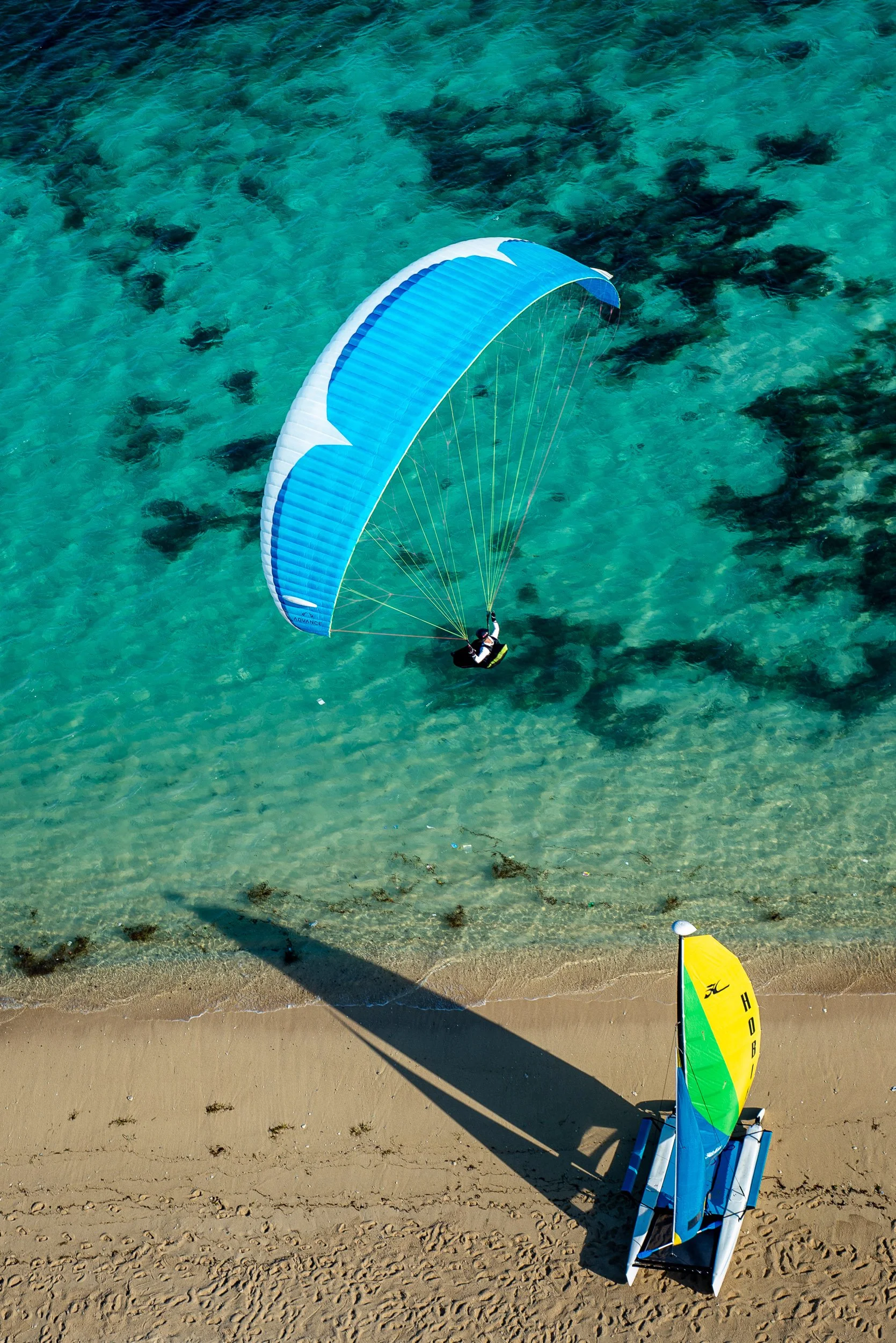 Paraglider flying over clear turquoise water with visible underwater rocks, sandy beach with footprints, and a colorful windsurfing sail on the shore.