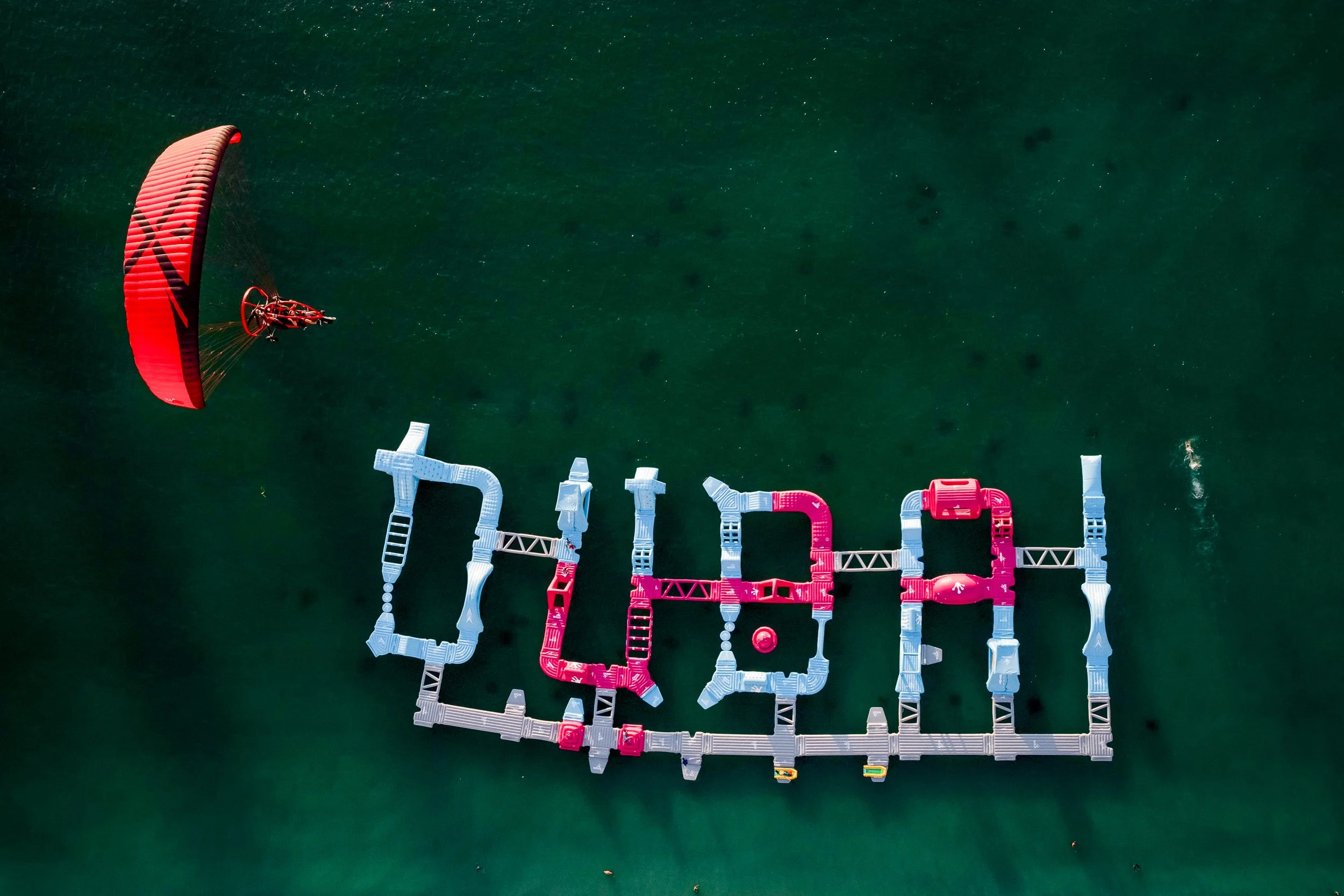 An aerial view of a green water body with a person flying a red kite and a floating letter display that spells 'dob'.