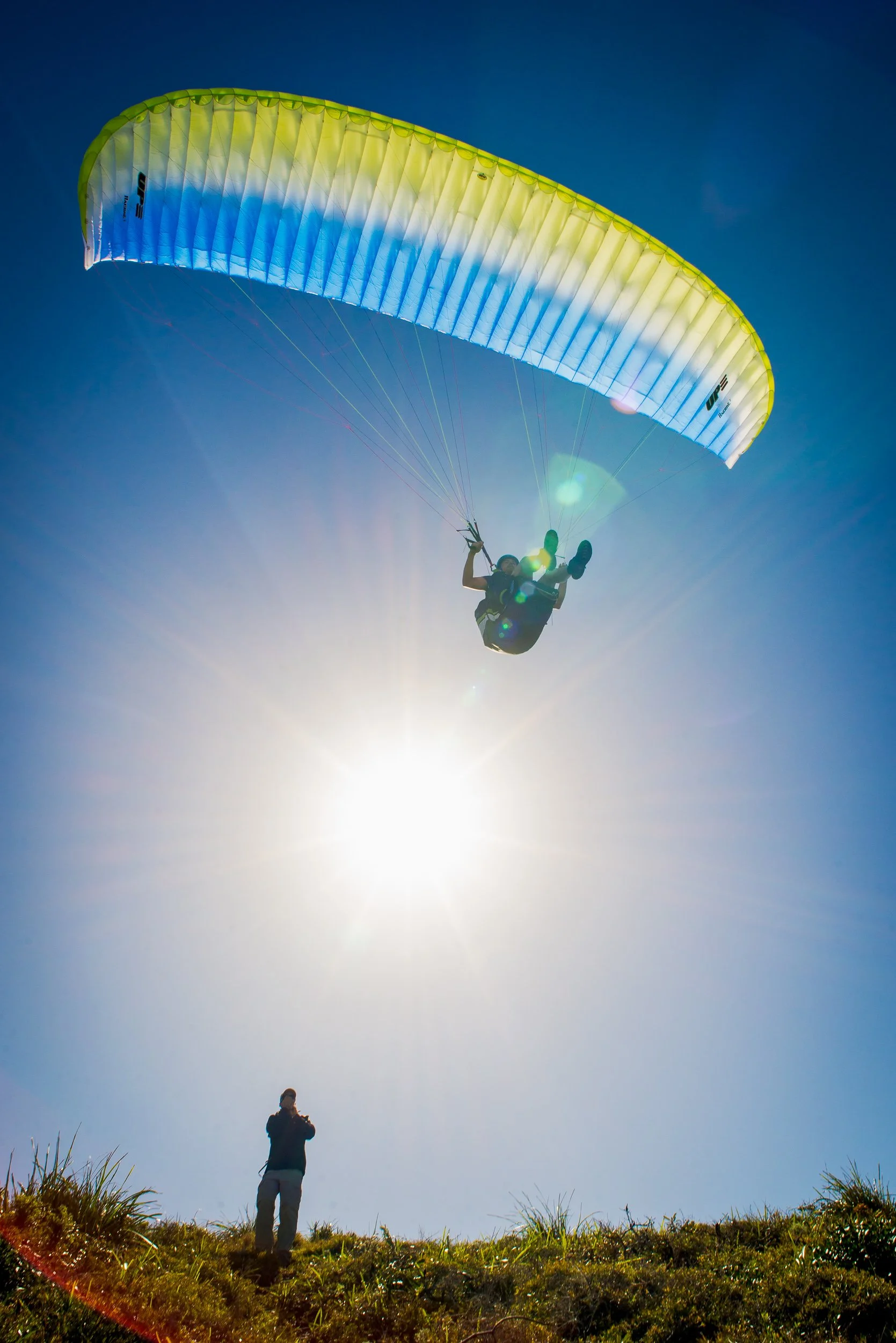 A person flying a blue and yellow paraglider in the sky, with another person standing on the ground watching. Bright sun shining, blue sky with no clouds.