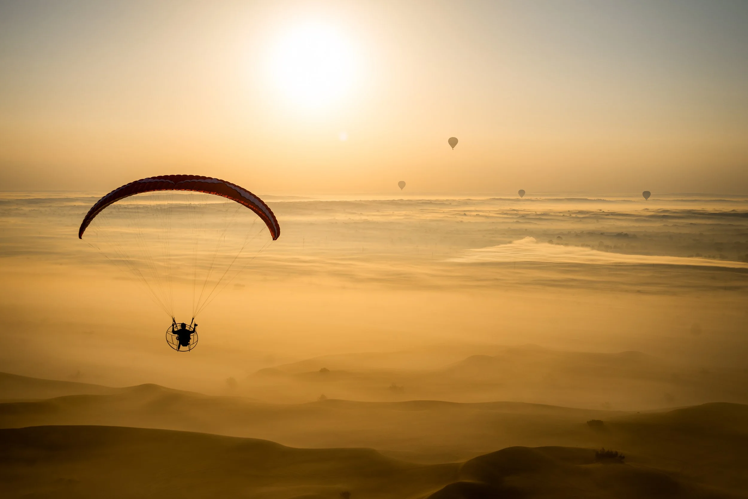 A person in a powered parachute flying over sand dunes during sunrise or sunset, with several hot air balloons in the sky.