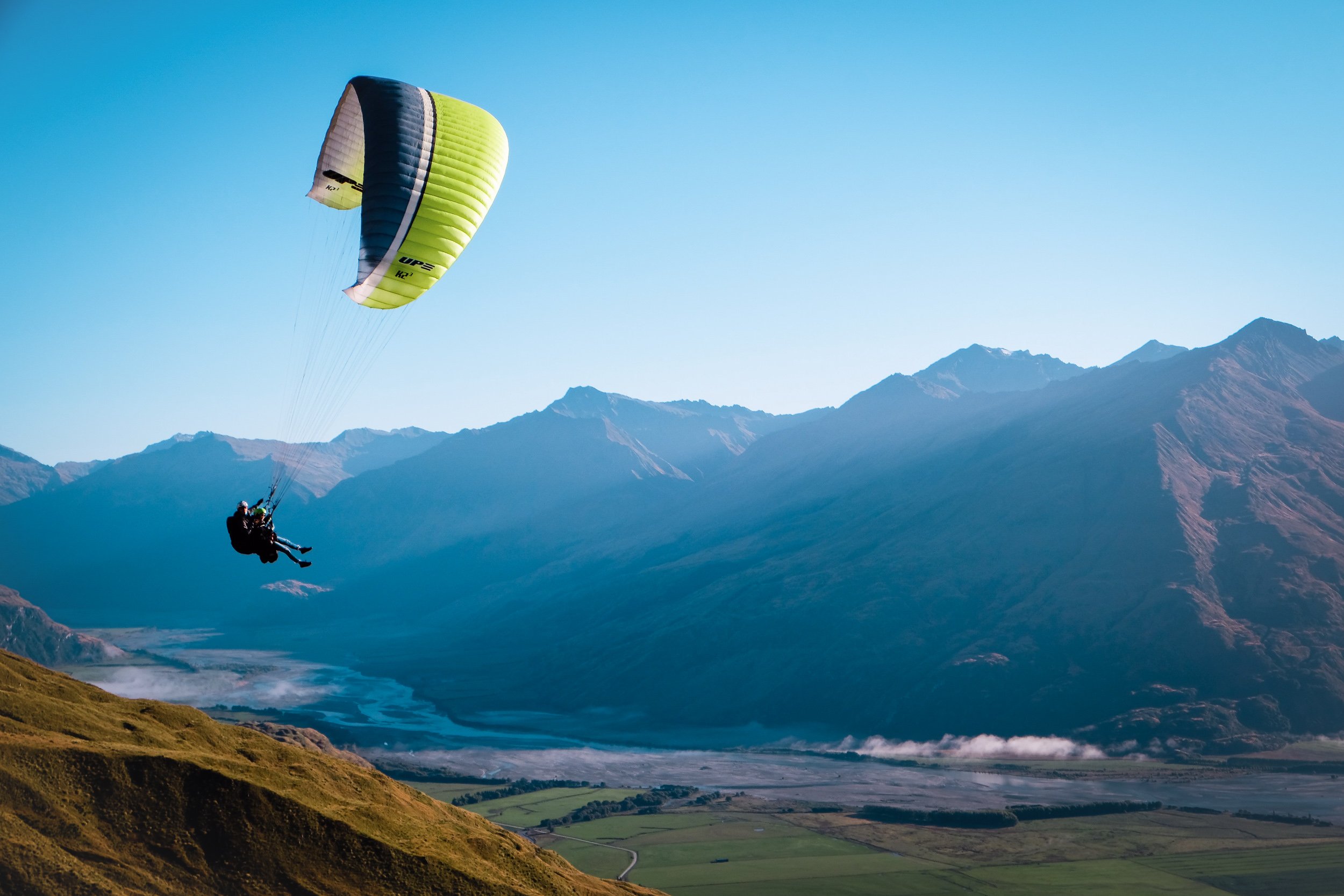 Two people paragliding over a mountainous landscape with a lake and green fields.