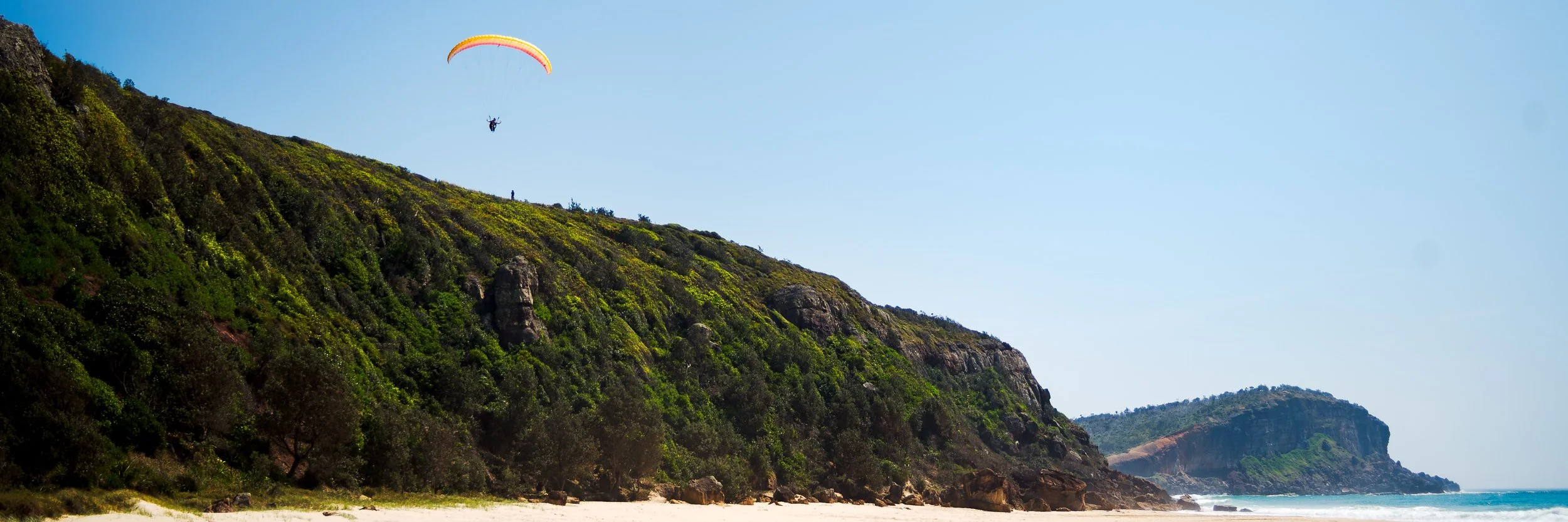 Paraglider flying over a green hillside at the beach with cliffs and the ocean in the background.