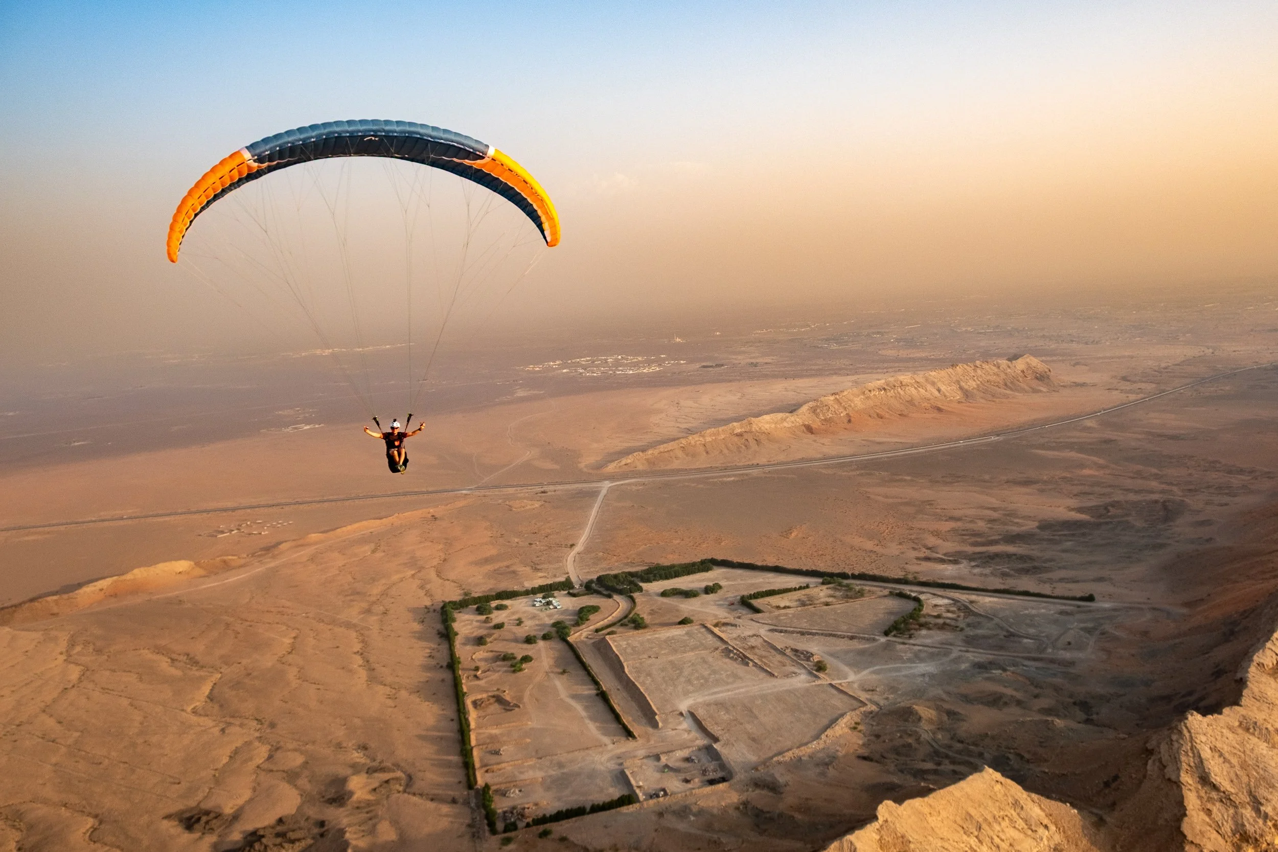 Person paragliding over a desert landscape during sunset with mountains in the distance.