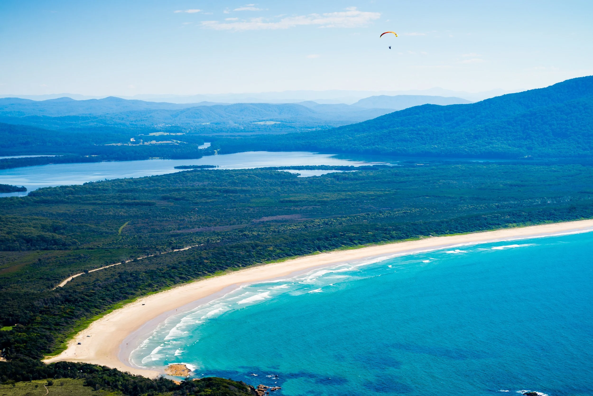 Aerial view of a coastal landscape featuring a sandy beach, blue ocean waves, lush green forests, and distant mountains under a clear sky with a paraglider in the sky.