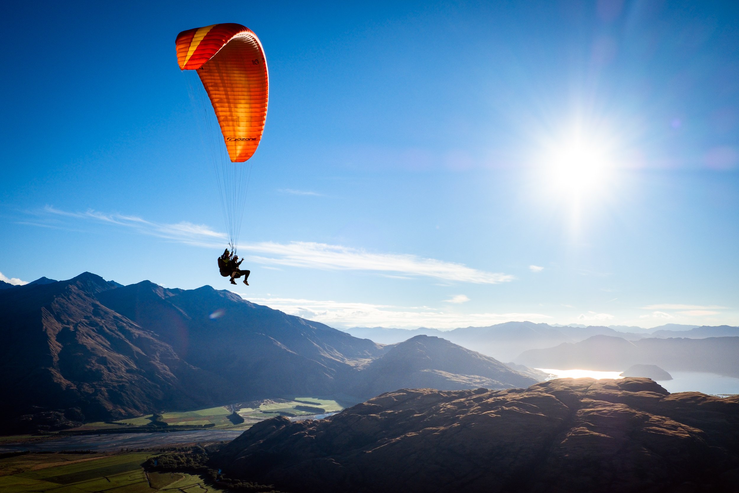A person paragliding over a mountainous landscape with a blue sky and the sun shining brightly.