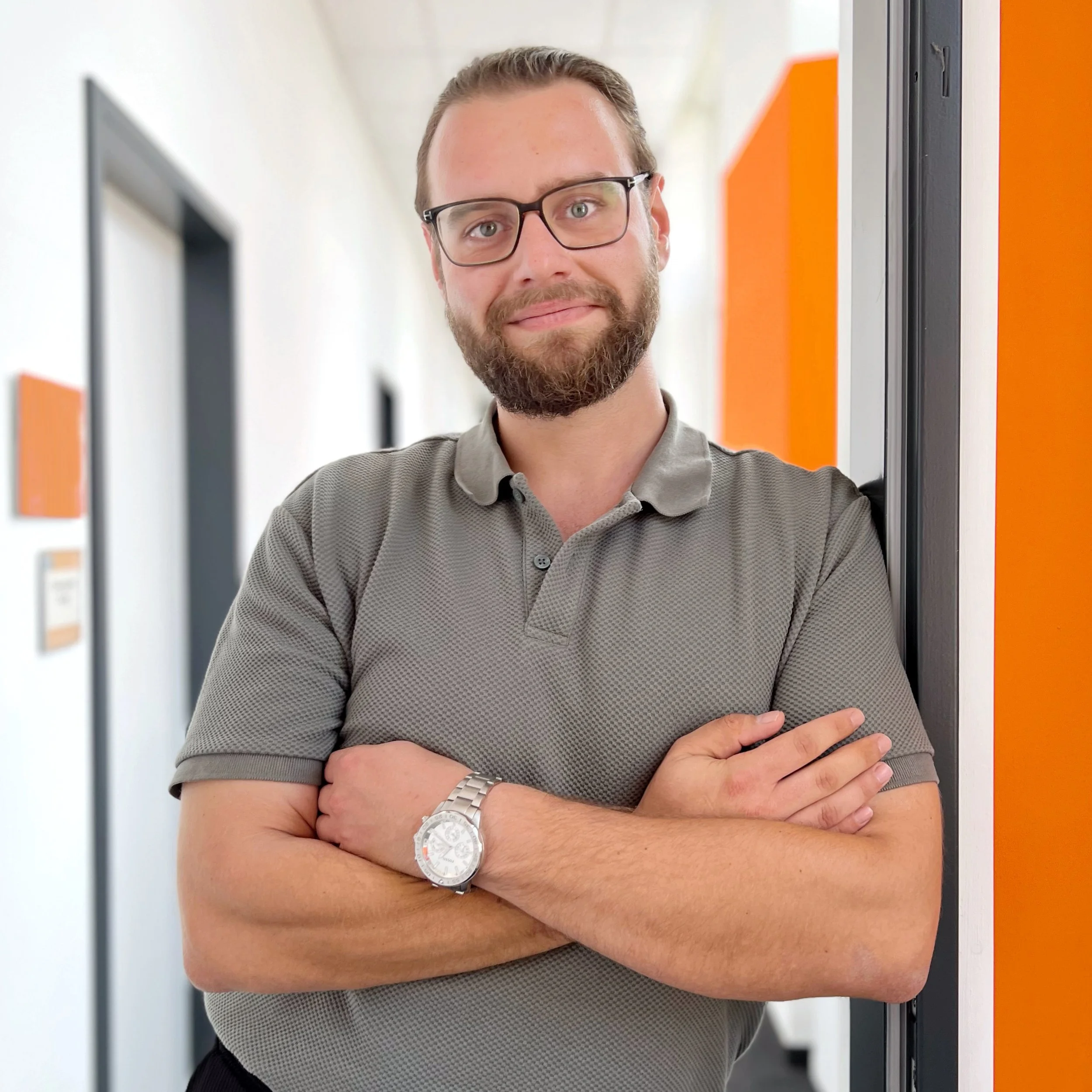 Jiga Consulting - Ein junger Mann mit Brille, Vollbart und Kurzhaar, trägt ein graues Hemd und eine Armbanduhr, steht vor orangefarbenen Schranktüren in einem modernen Büro.