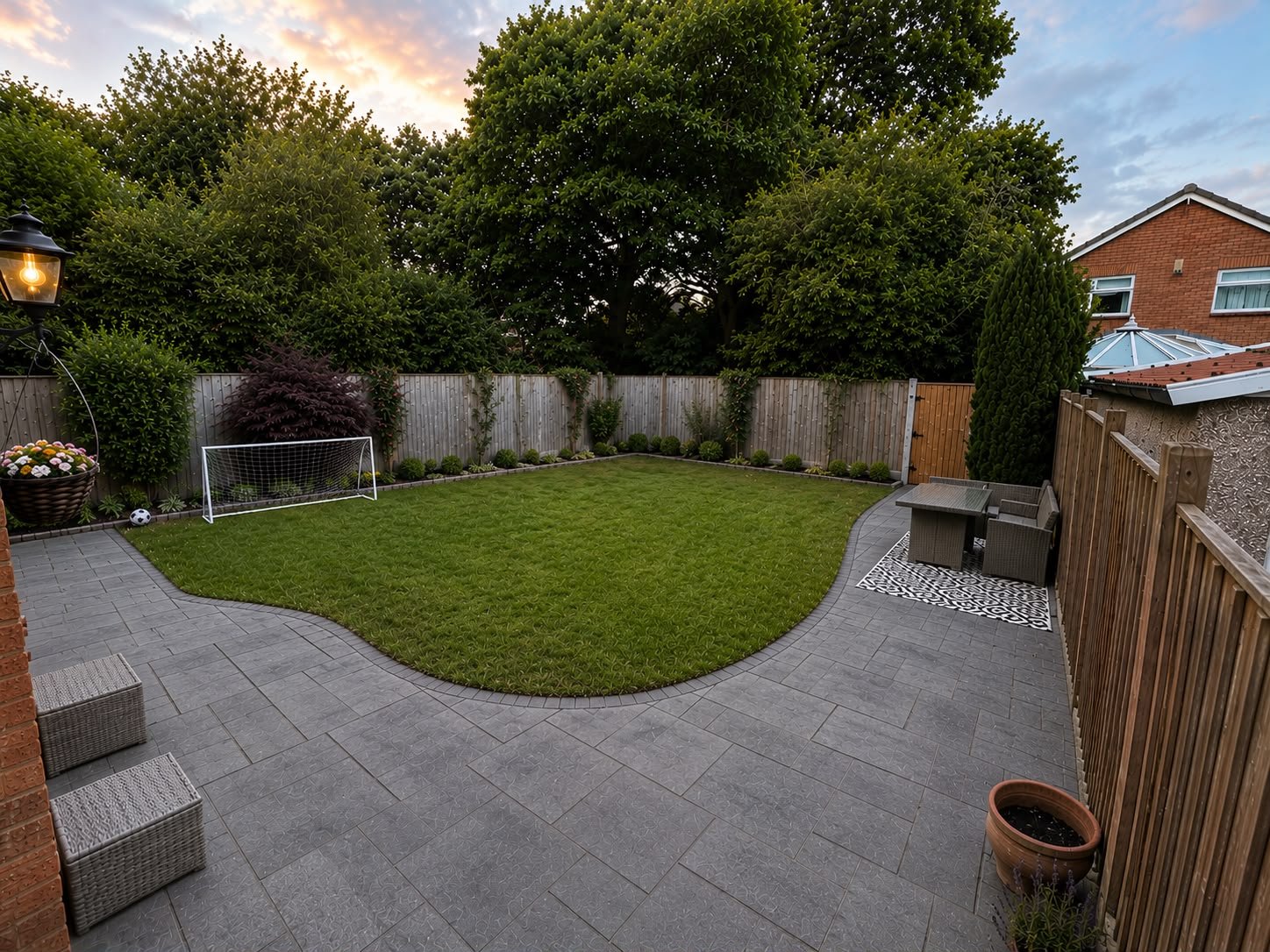 A backyard with a small soccer goal, a lush green lawn bordered by shrubs, a brick and wooden fence, outdoor furniture including a table and chairs, and decorative paving stones. A lamp hangs from a pole near flower pots, and large trees are visible beyond the fence.