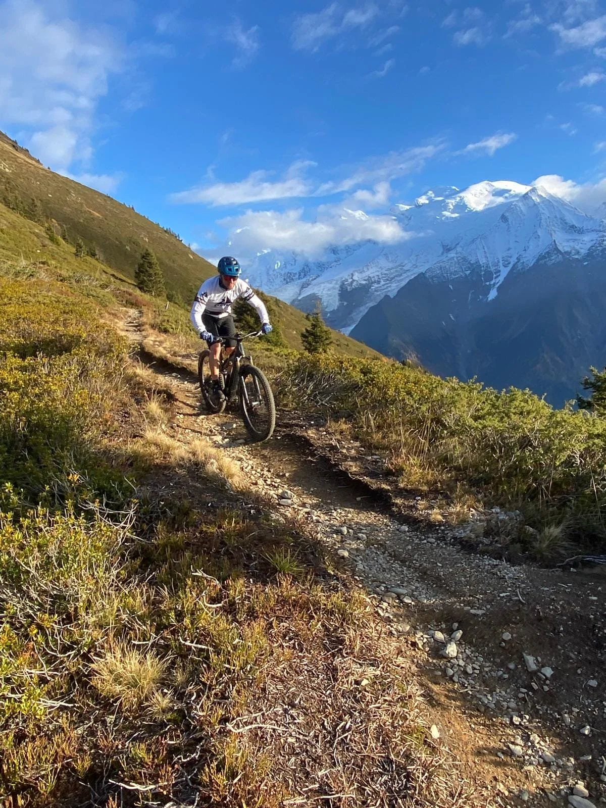 A mountain biker riding on a rugged trail in a mountainous landscape with snow-capped peaks in the background.