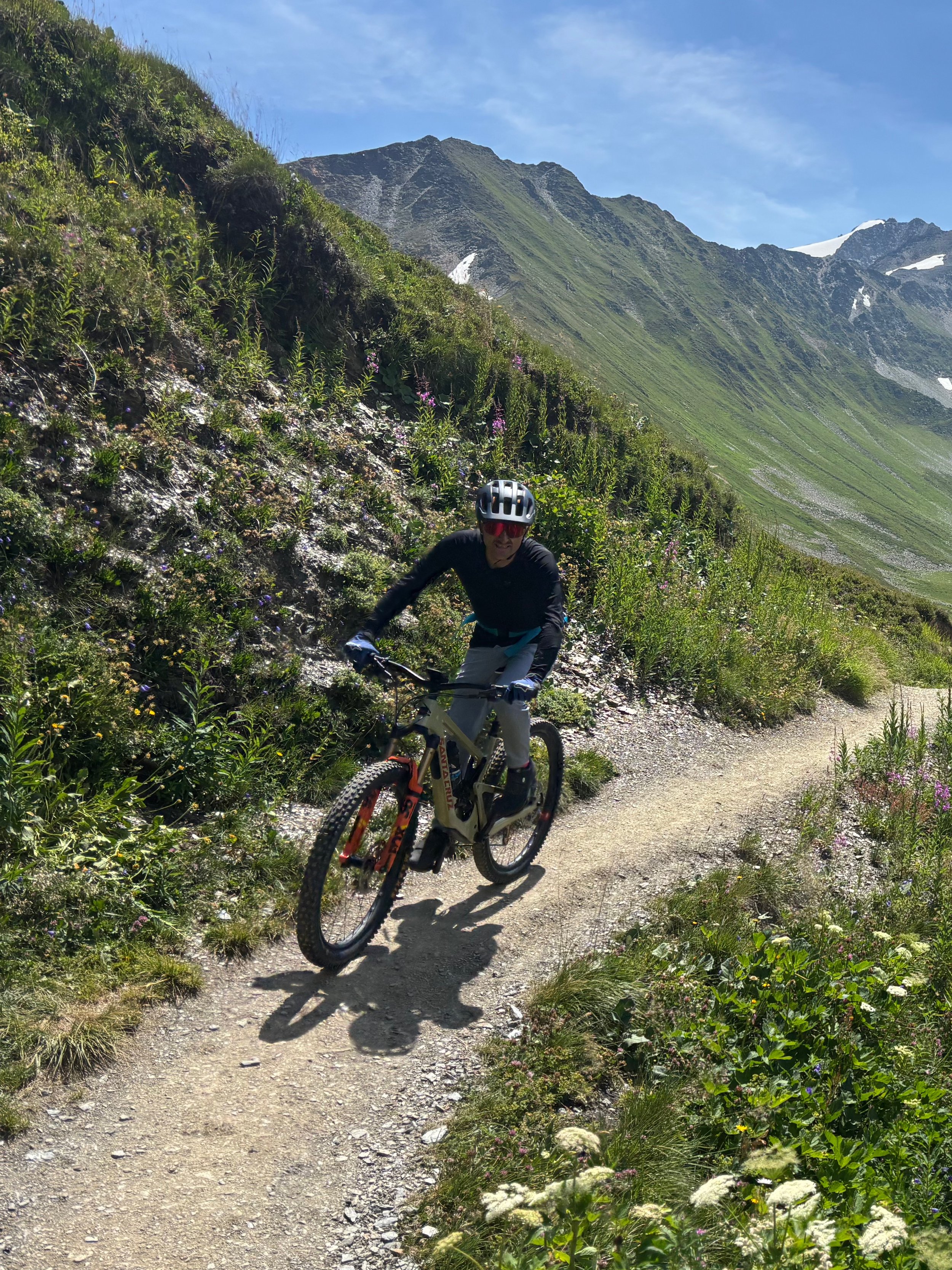 A person mountain biking on a dirt trail in a mountainous area with green vegetation, snow patches on the peaks, and a clear blue sky.