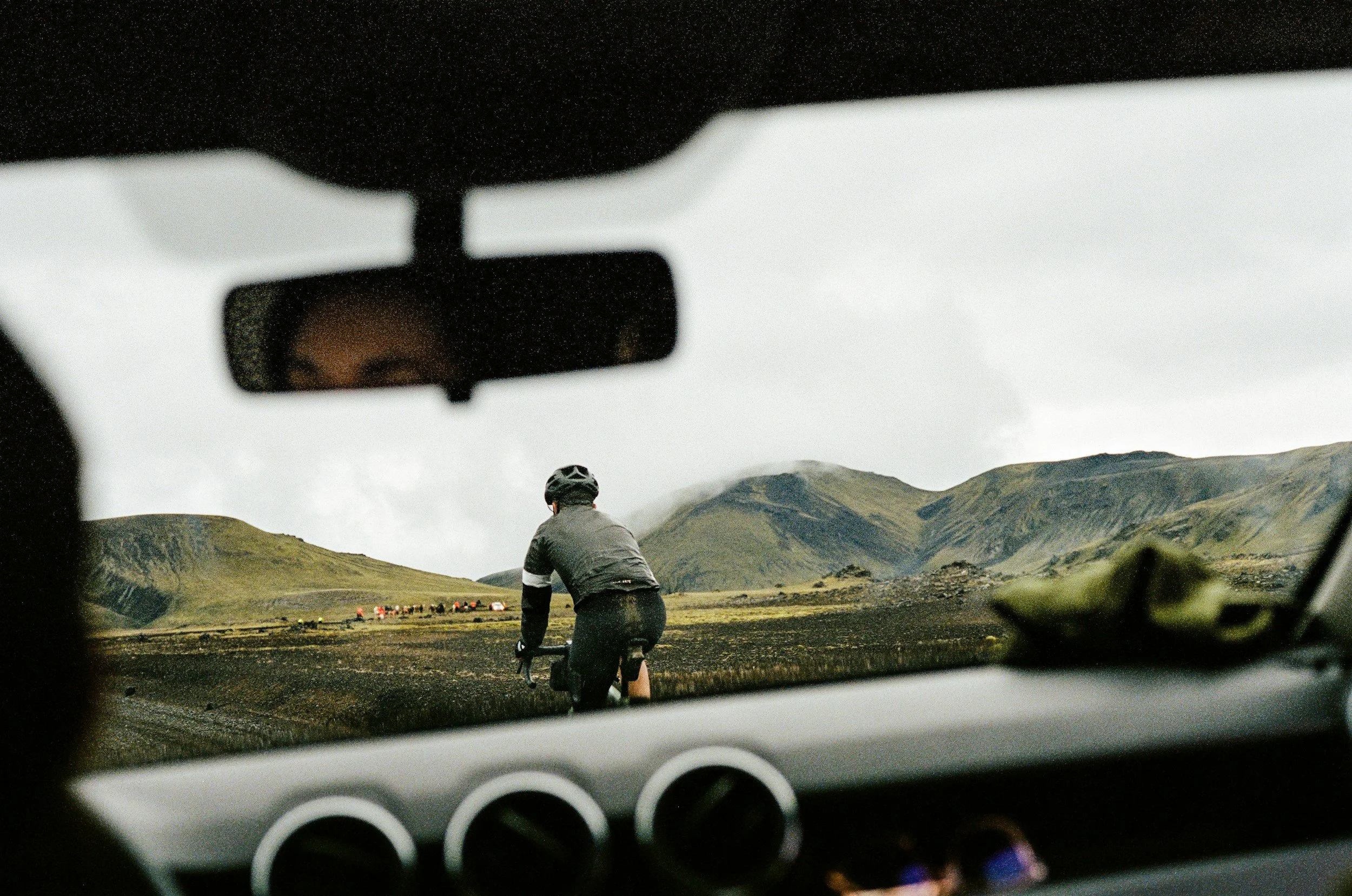 A person riding a bicycle on a dirt path in a mountainous landscape, viewed from inside a car with the rearview mirror and dashboard in the foreground.