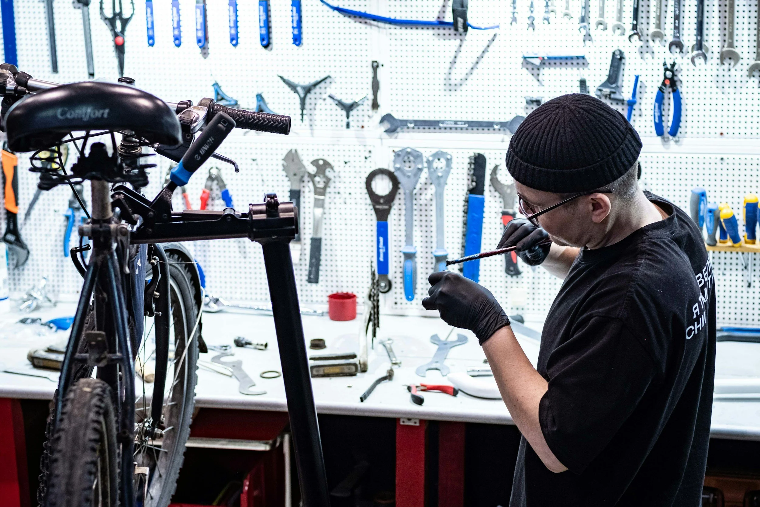 A person wearing a black beanie, glasses, and black gloves working on a bicycle in a workshop with tools hanging on a pegboard in the background.