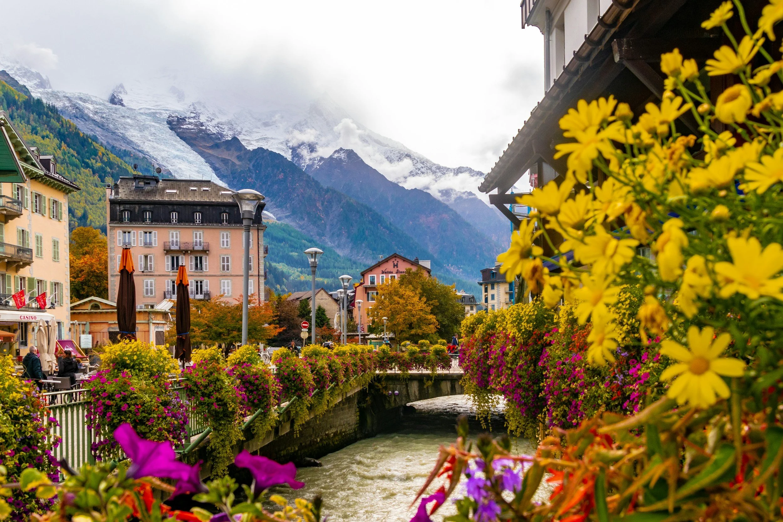 A picturesque town with colorful buildings and vibrant flower beds along a river, snow-capped mountains in the background, and partially cloudy skies.