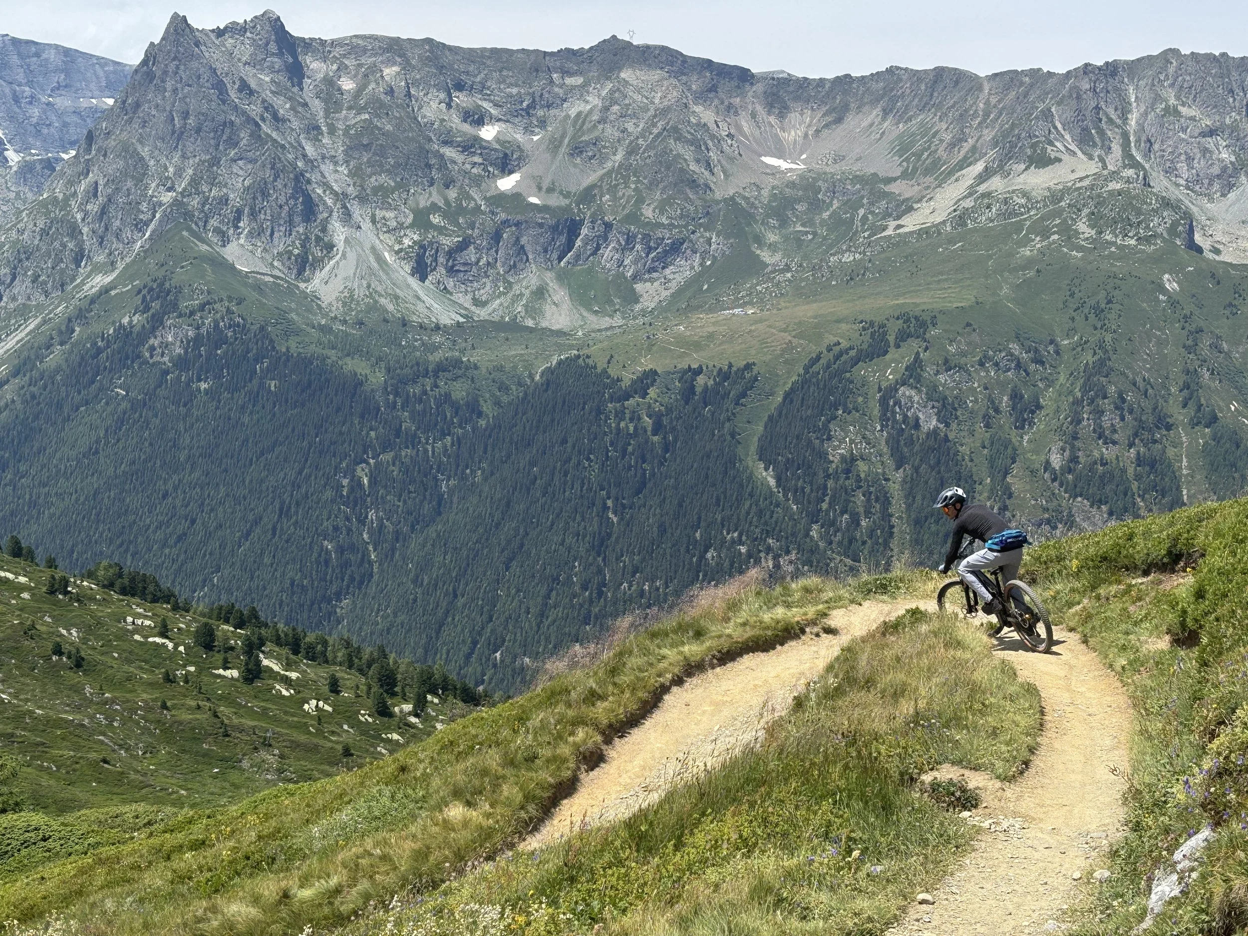 A person mountain biking on a dirt trail in a mountainous landscape with green slopes and rocky peaks in the background.