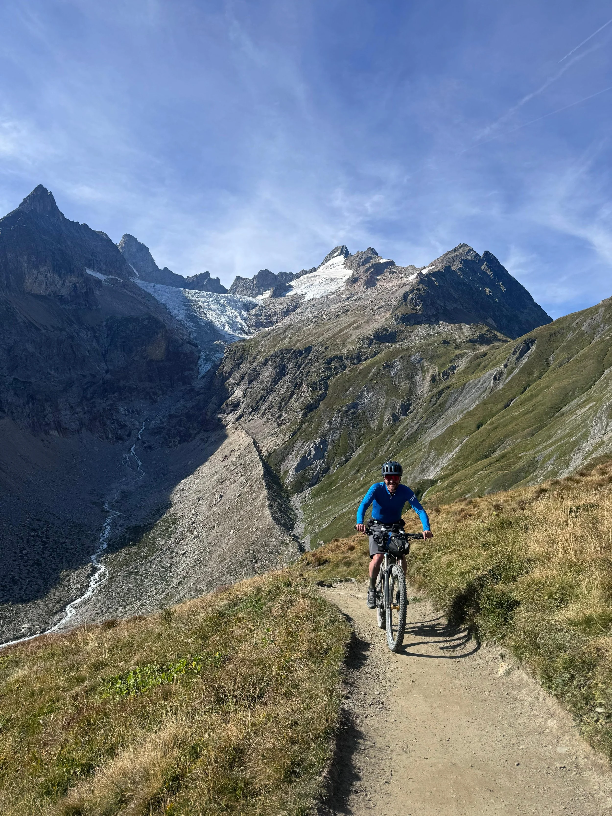 A cyclist riding on a narrow mountain trail surrounded by grassy slopes with towering mountains and snow patches in the background under a blue sky.