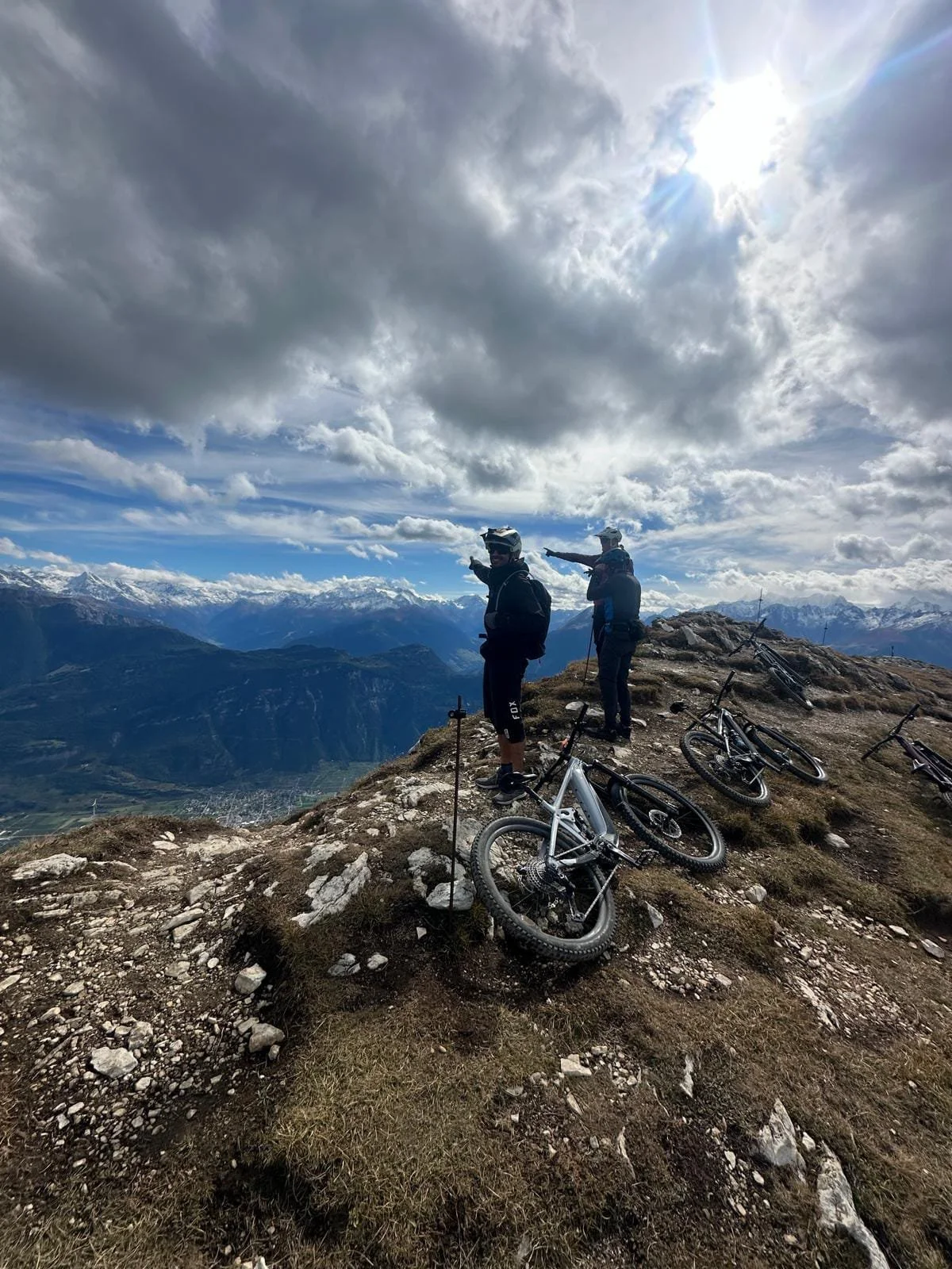 Two mountain bikers wearing helmets and backpacks standing on a rocky mountain peak with their bikes lying on the ground nearby, overlooking a vast scenic view of mountains and clouds in the sky.